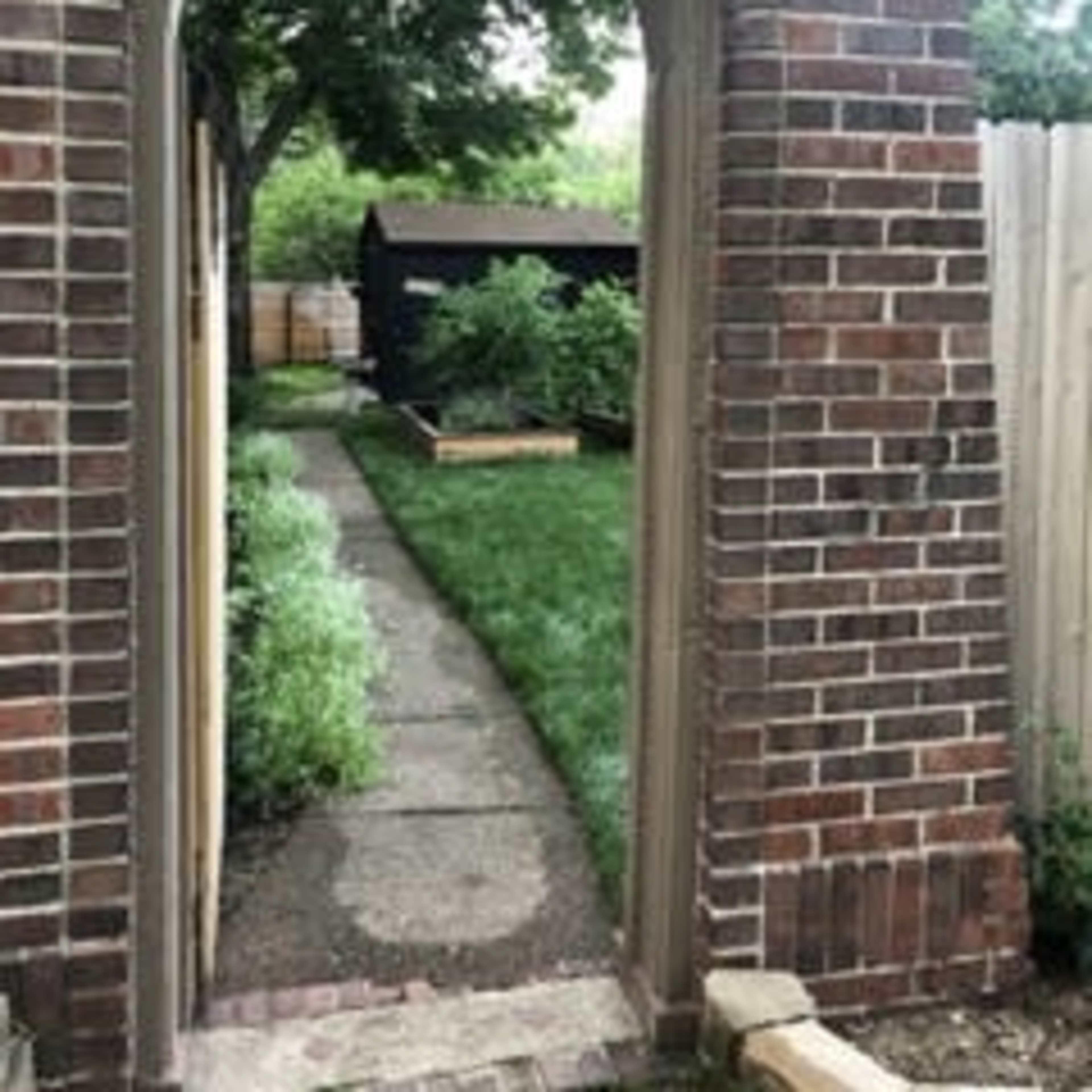 An archway made of brick frames a stone path leading to a garden shed behind a grassy area.