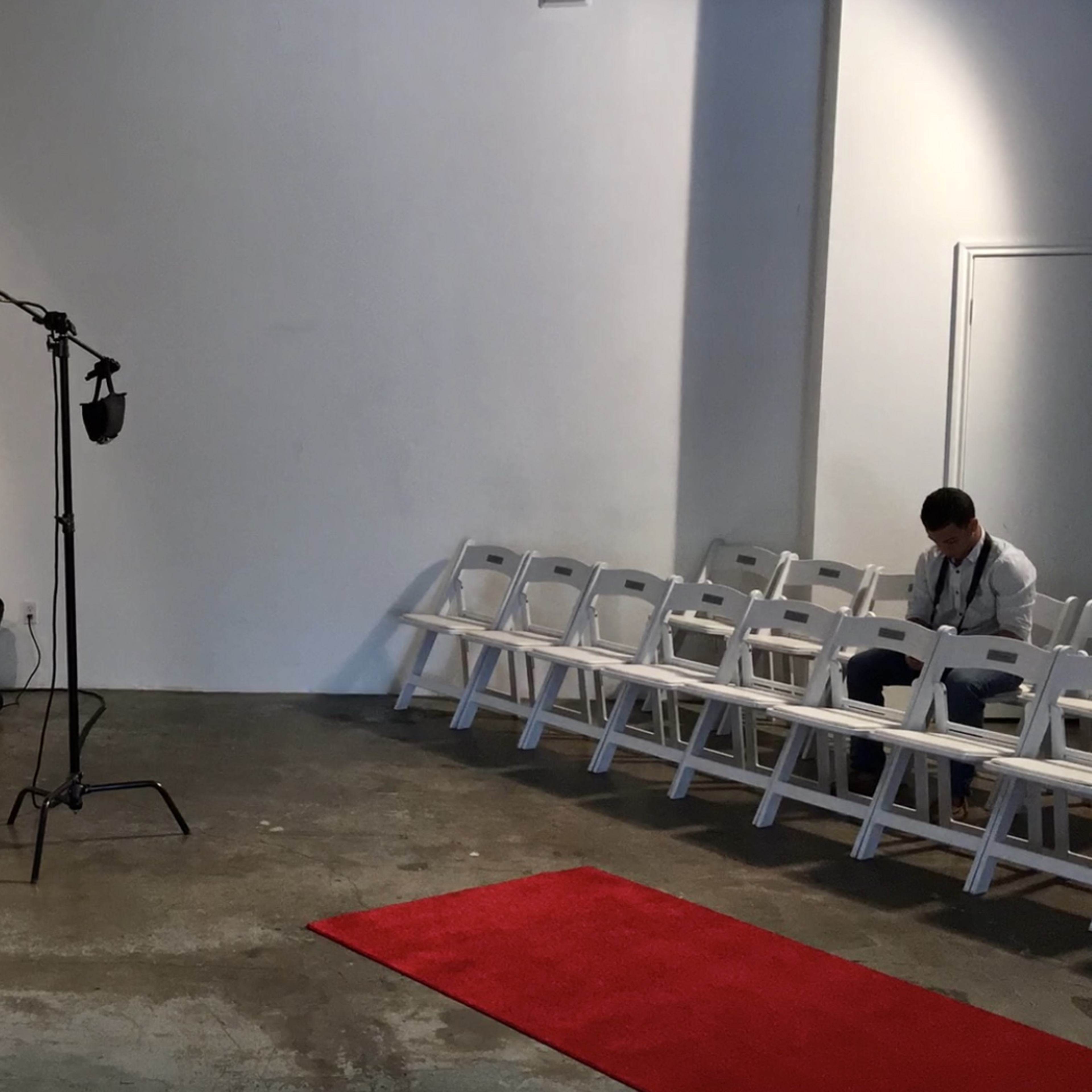 A person stands on a small stage with two lights pointed at them, while several empty white chairs are arranged in front of the set.