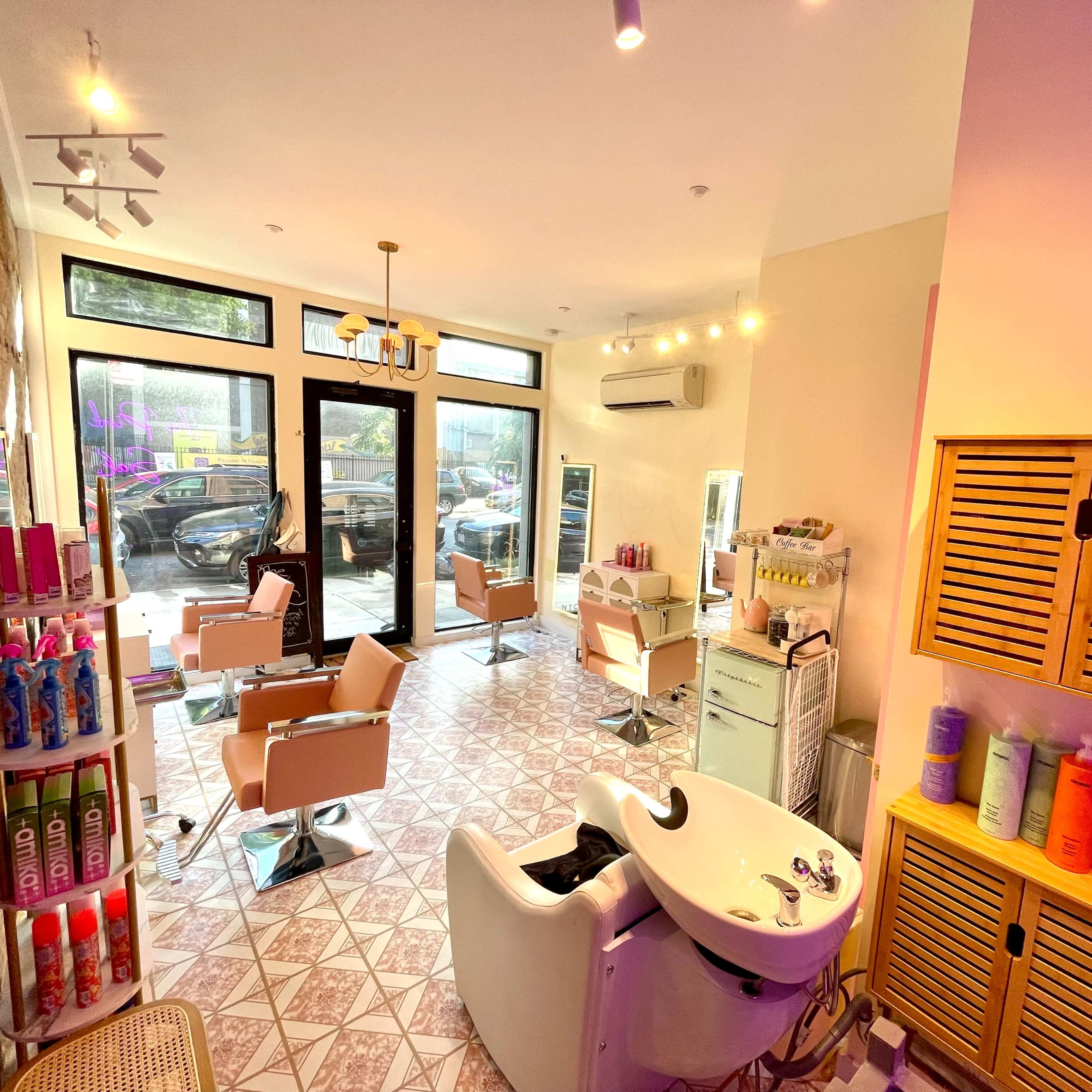 A modern hair salon with several styling chairs, a shampoo station, and shelves stocked with hair products, all framed by large windows allowing natural light to enter.