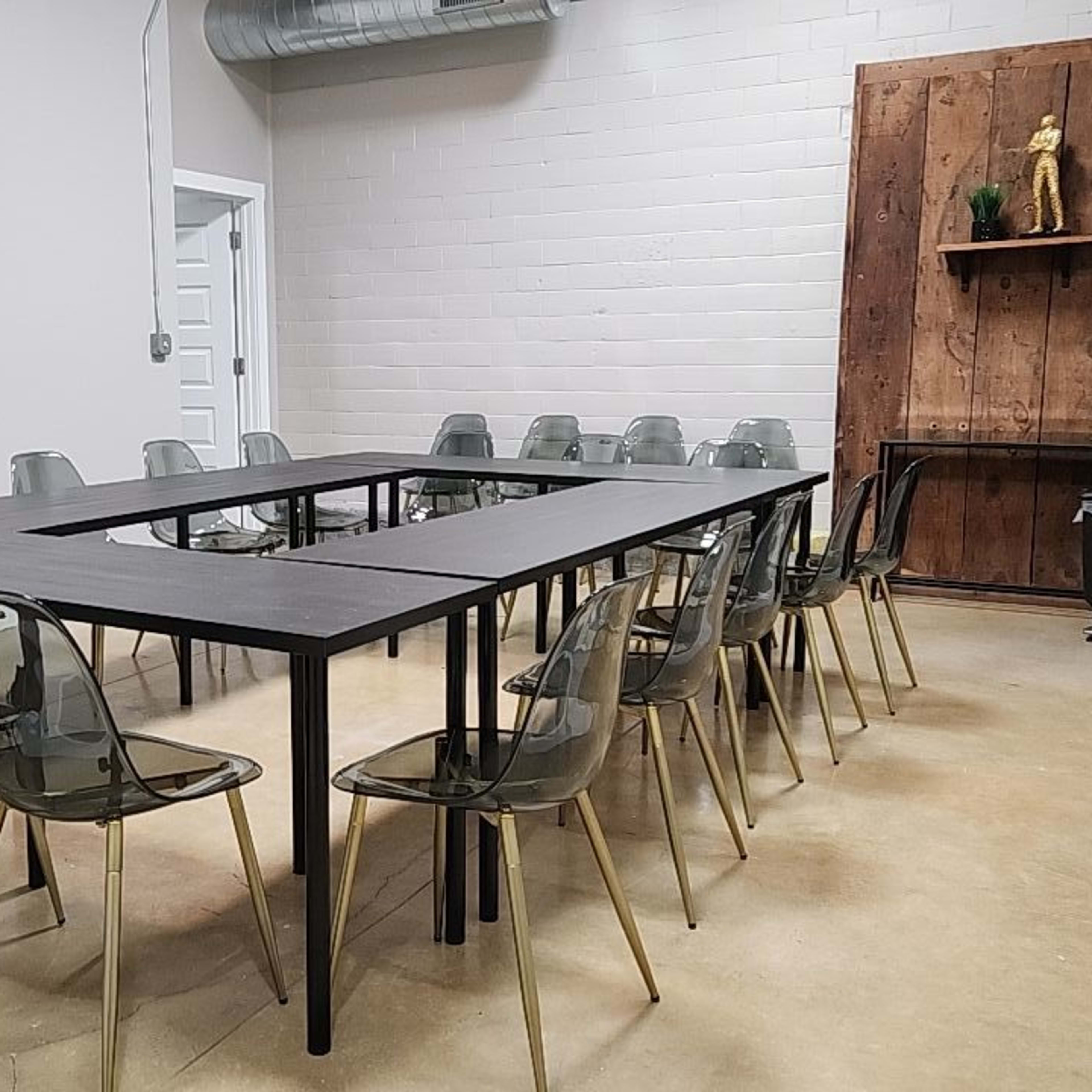 A conference room features a rectangular table surrounded by transparent chairs, with wooden paneling and a plant displayed against the wall.