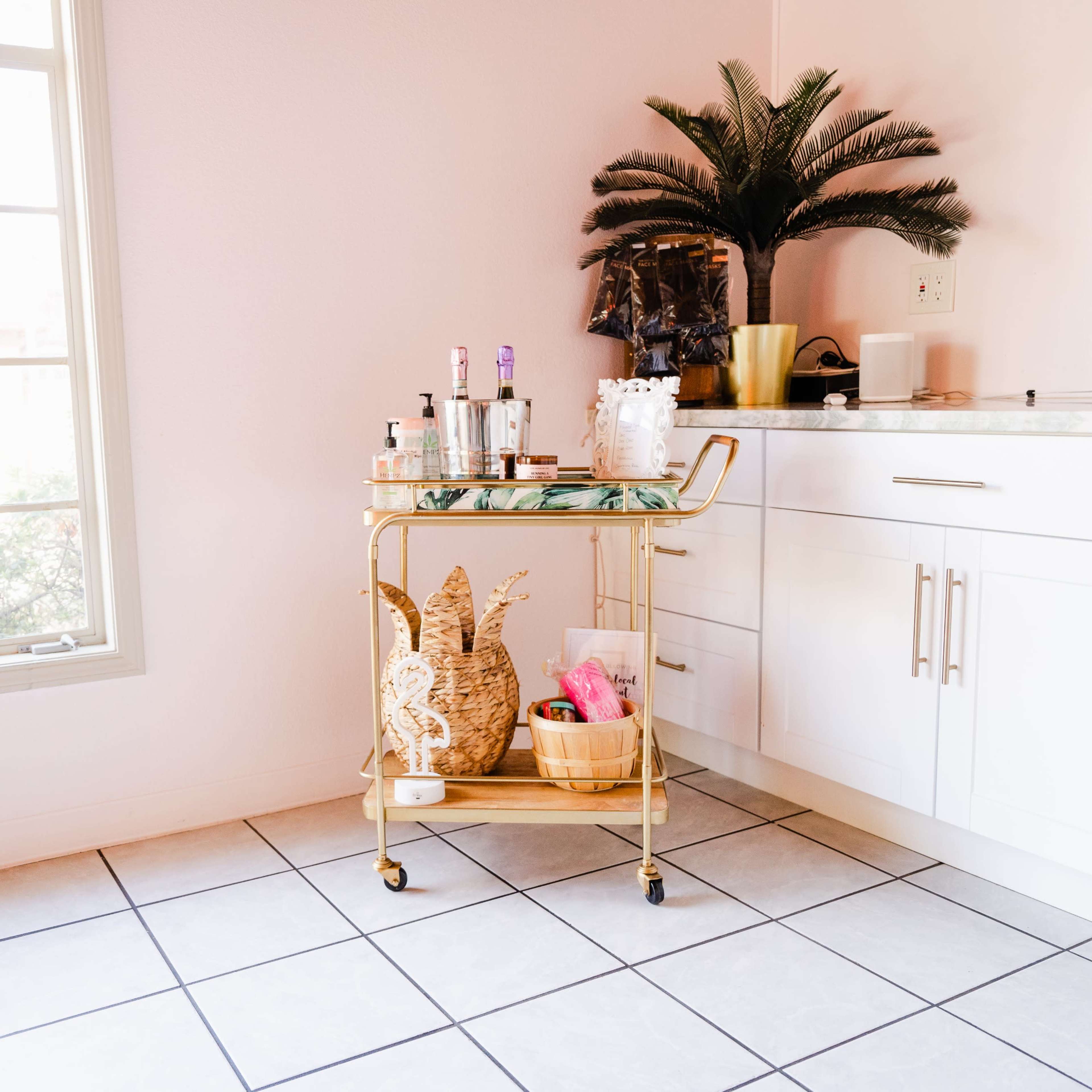 A gold bar cart with various bottles and decorative items is positioned next to a white cabinet beside a window.