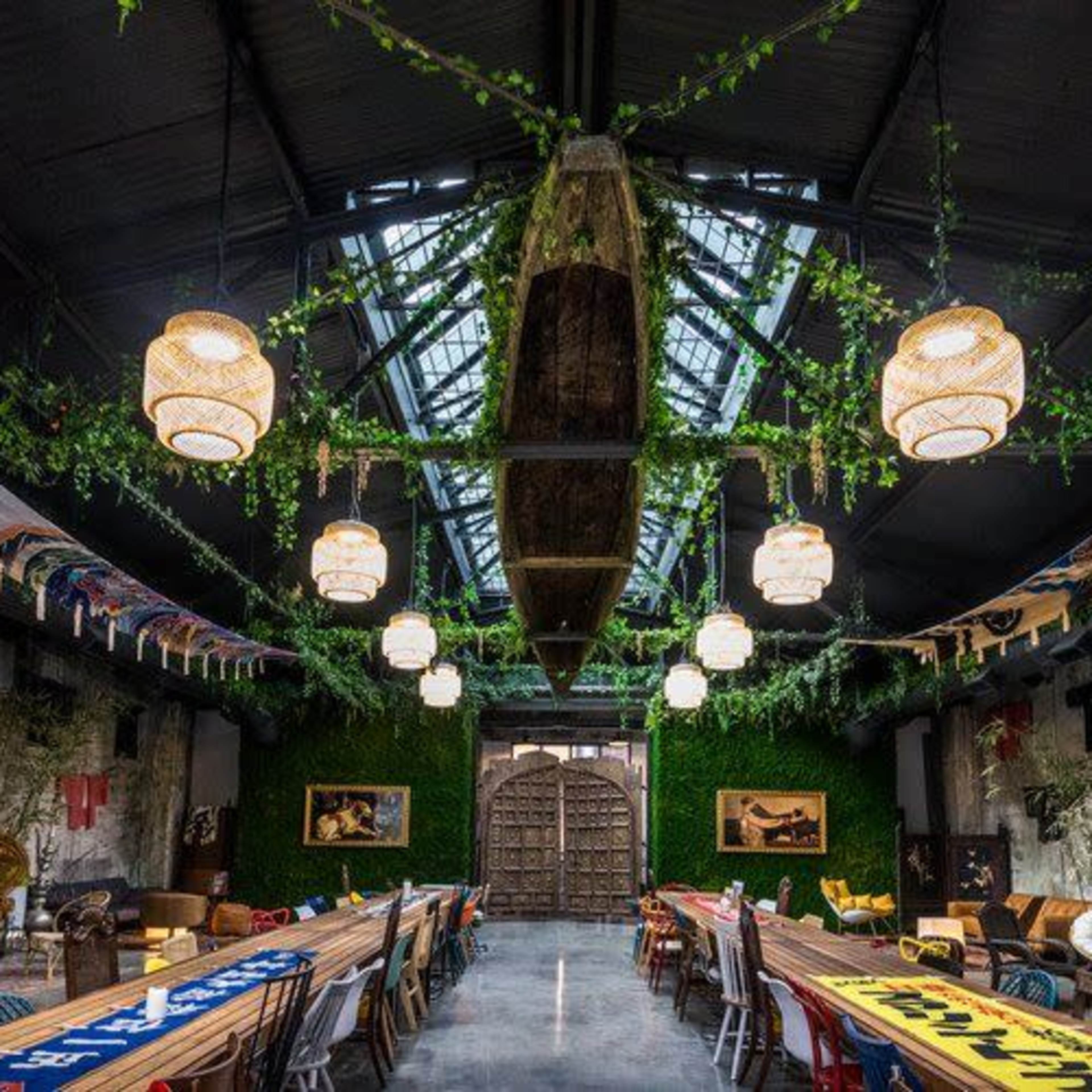 An industrial-style hall with a high ceiling, adorned with plants and banners, featuring long tables arranged under pendant lights.