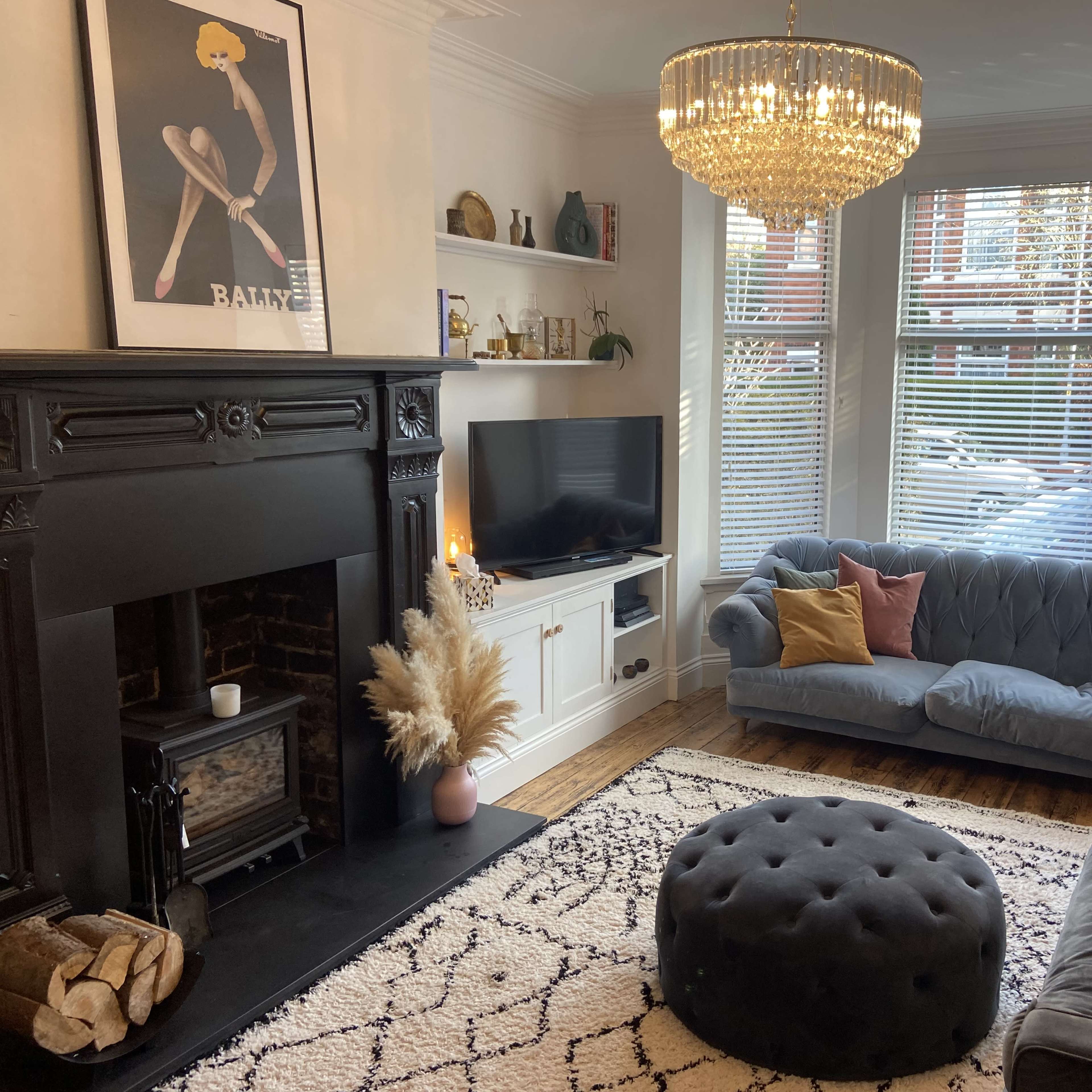 A stylish living room featuring a chandelier, a brick fireplace, a television, a blue velvet sofa, and a round ottoman on a patterned rug.