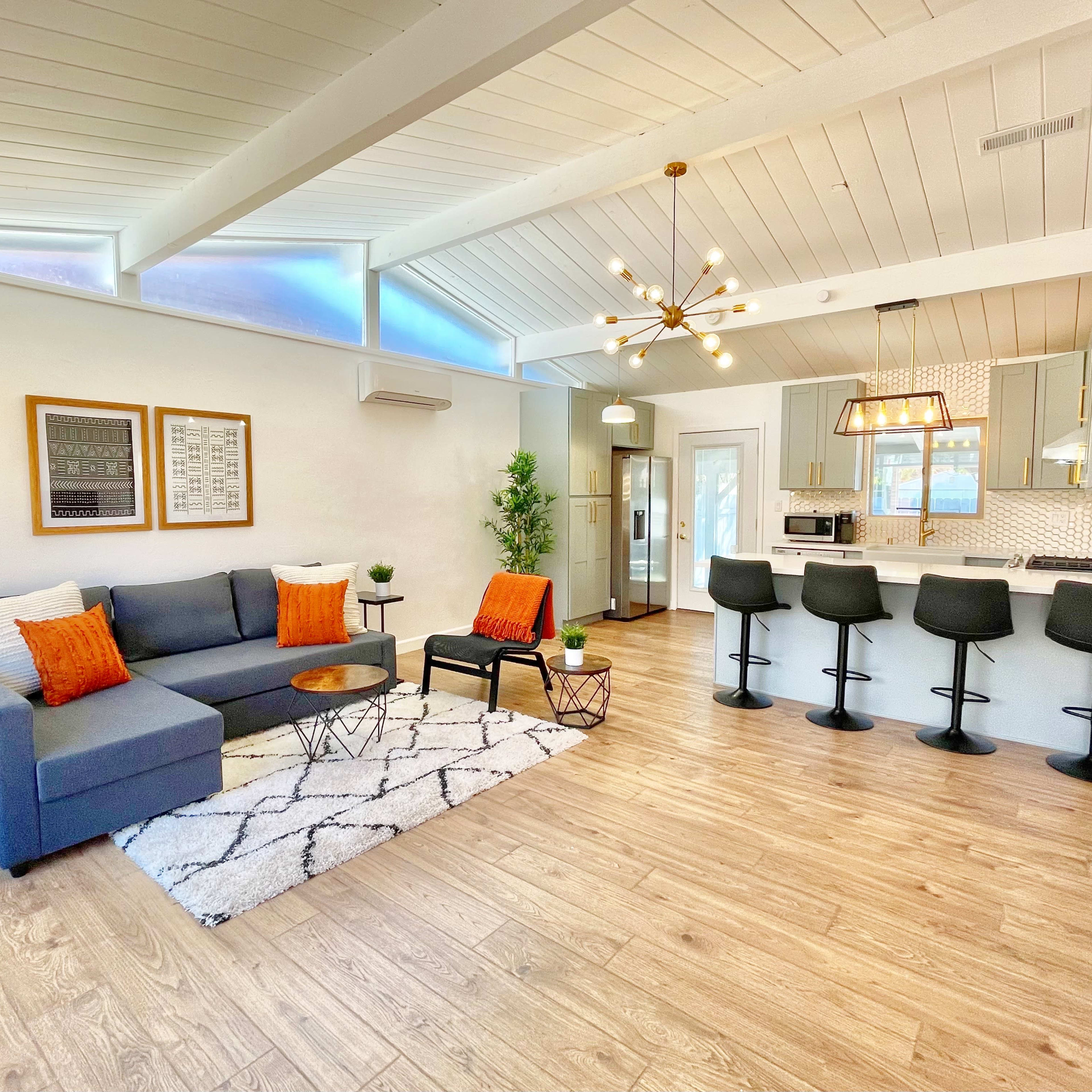 A modern living room with a sectional sofa, decorative pillows, a coffee table, and a kitchen area featuring bar stools and stainless steel appliances.