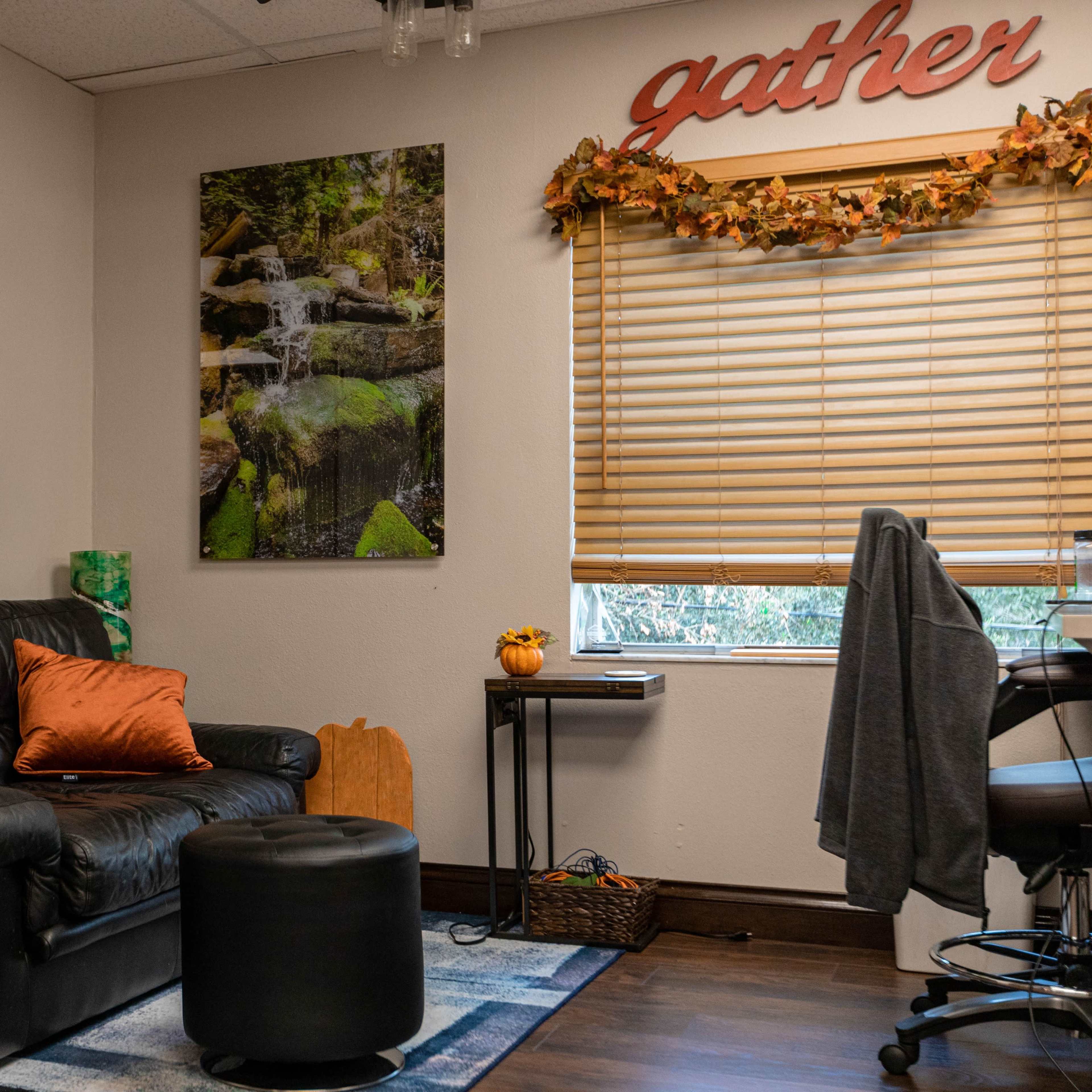 A small office space featuring a black leather couch, a round black ottoman, a desk with a chair, and nature-themed wall art above the couch.