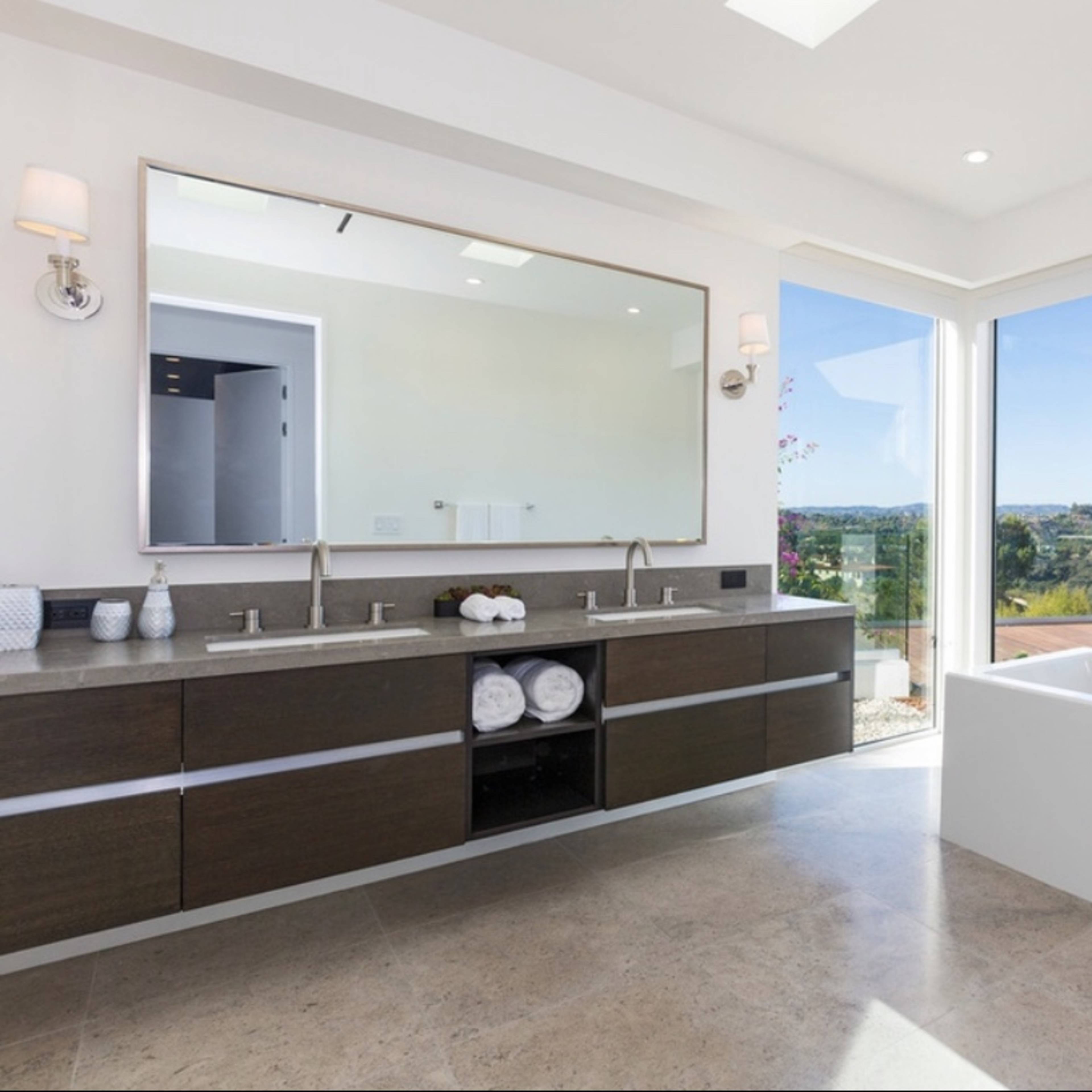 A modern bathroom featuring a double sink vanity, large mirror, a freestanding bathtub, and floor-to-ceiling windows with a view of the landscape.
