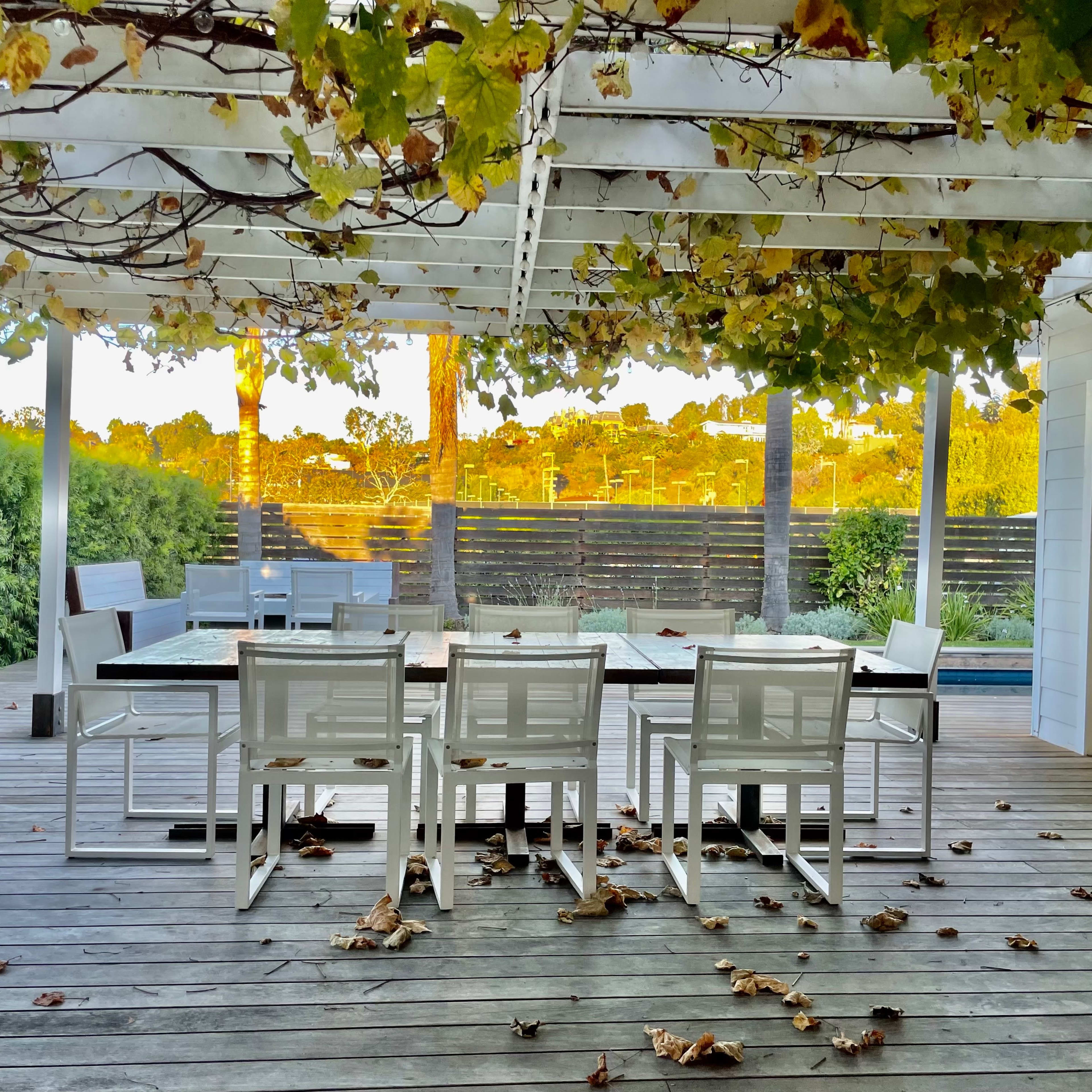 A spacious outdoor deck features a white wooden table with several chairs underneath a pergola covered in vines, surrounded by greenery and scattered leaves.