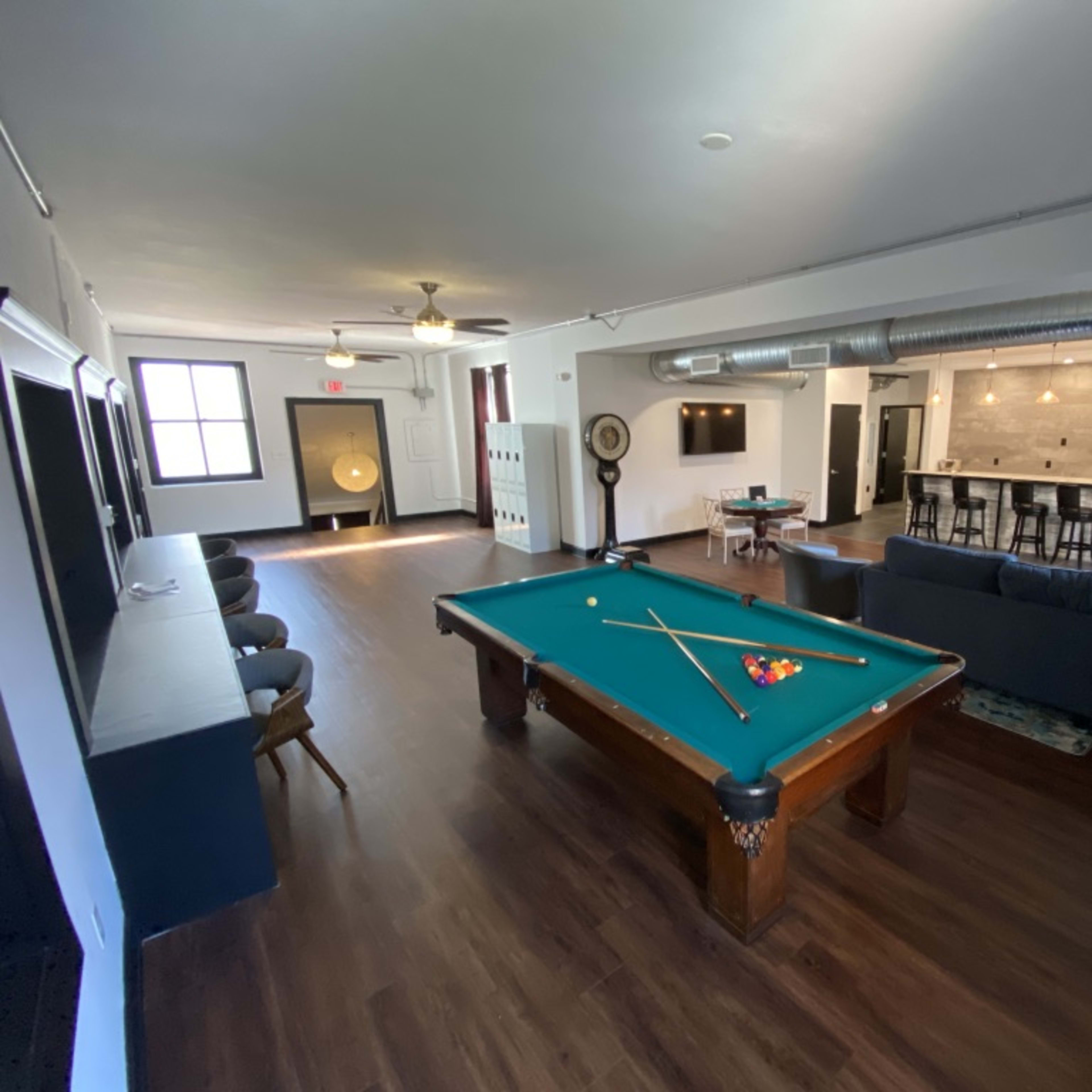 The room features a pool table in the foreground, with a seating area and a bar in the background.