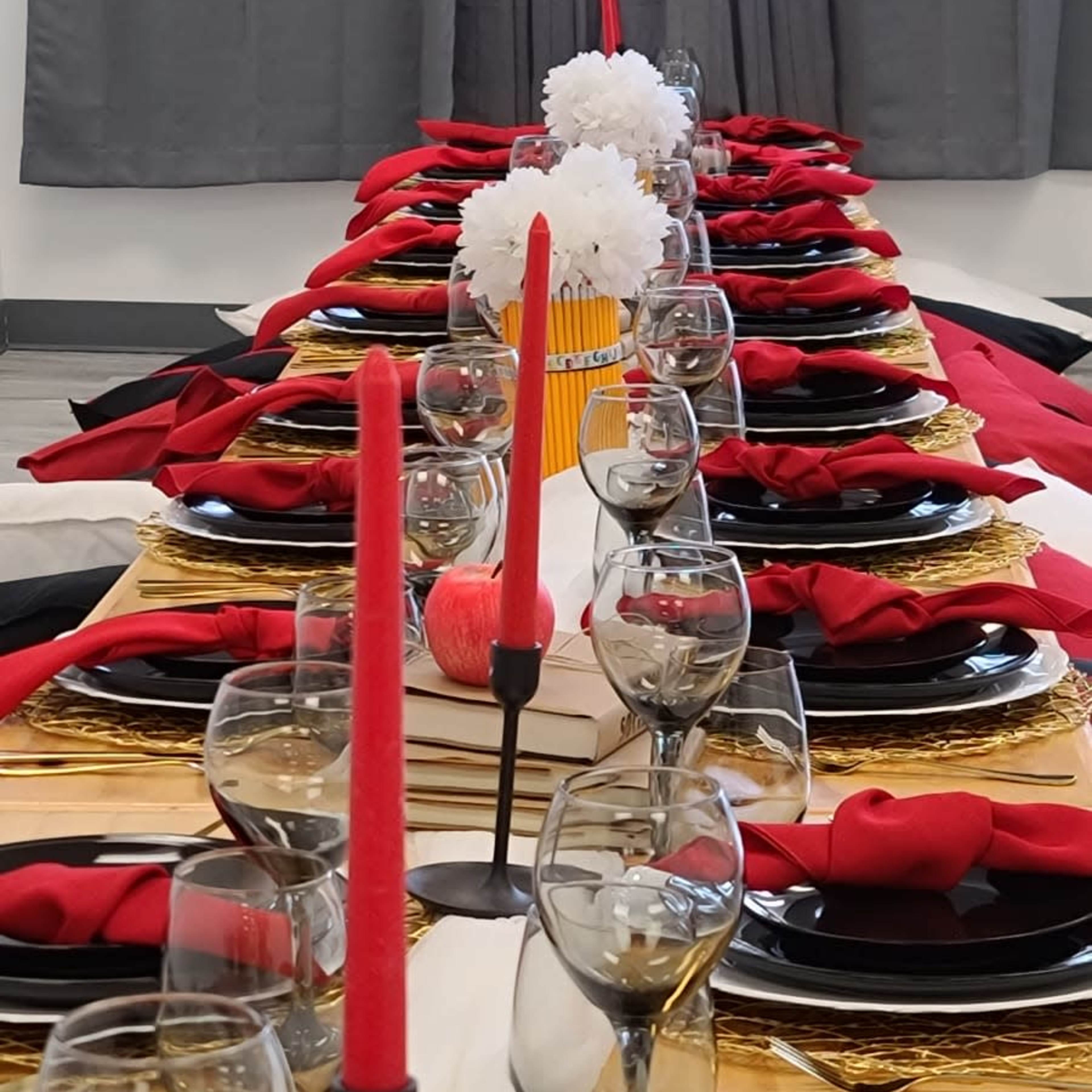 A long dining table is set with black plates, gold accents, red napkins, and red candles alongside glasses and a decorative centerpiece.