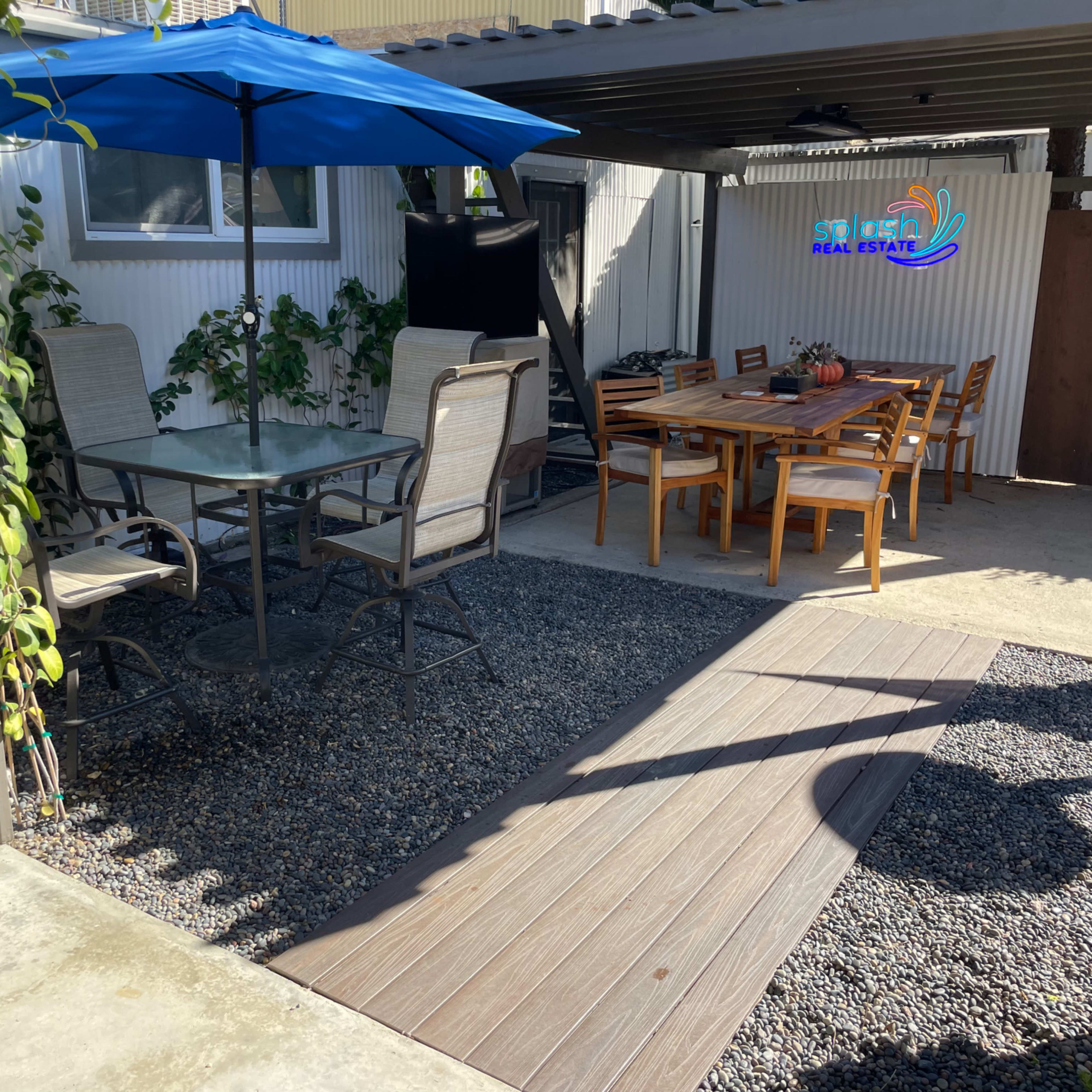 A backyard patio area with a dining table and chairs under an umbrella, alongside a gravel pathway and a wall displaying a sign for "Sierra Real Estate."