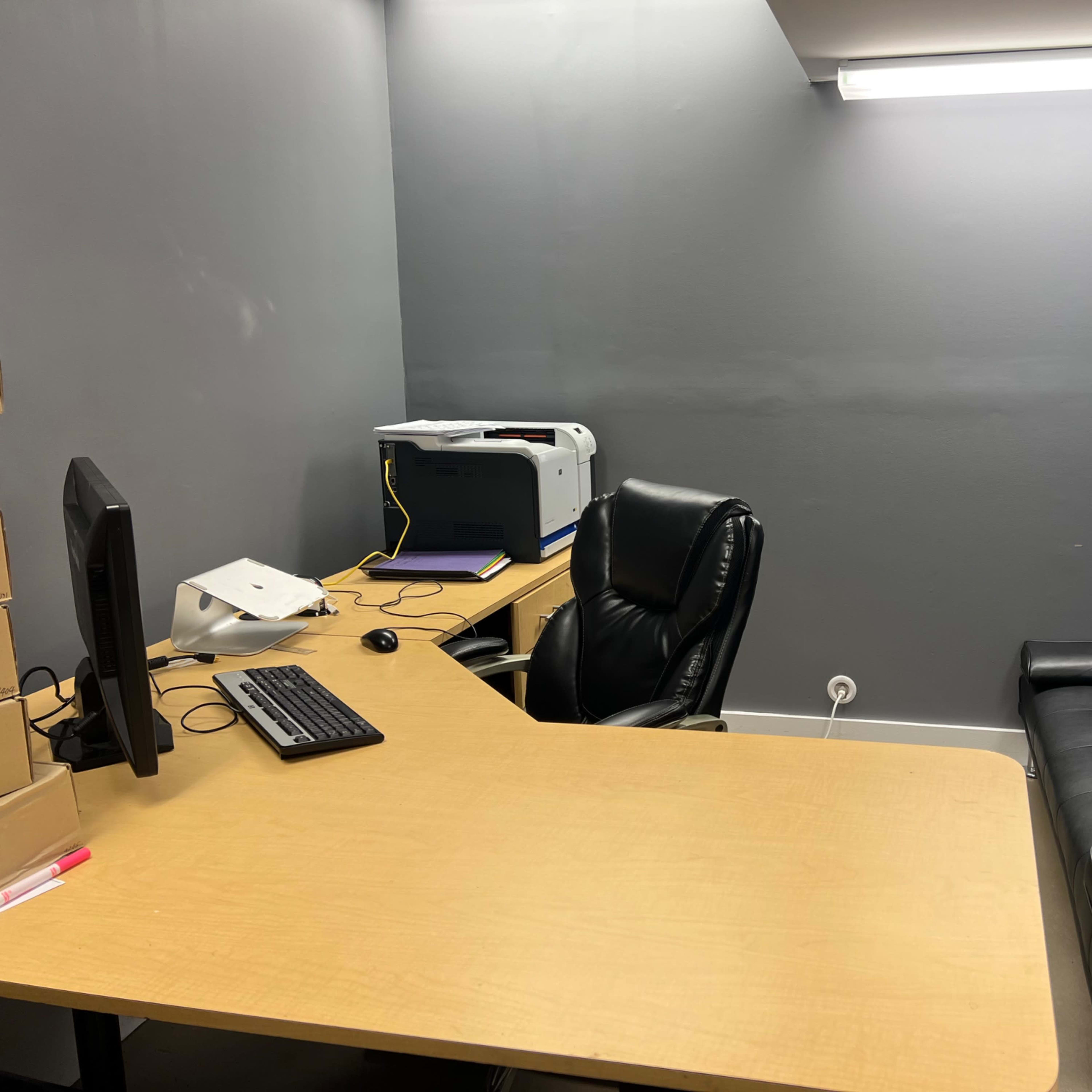 A small office space with a desk, a computer, a printer, a black leather chair, and a black sofa against the gray wall.