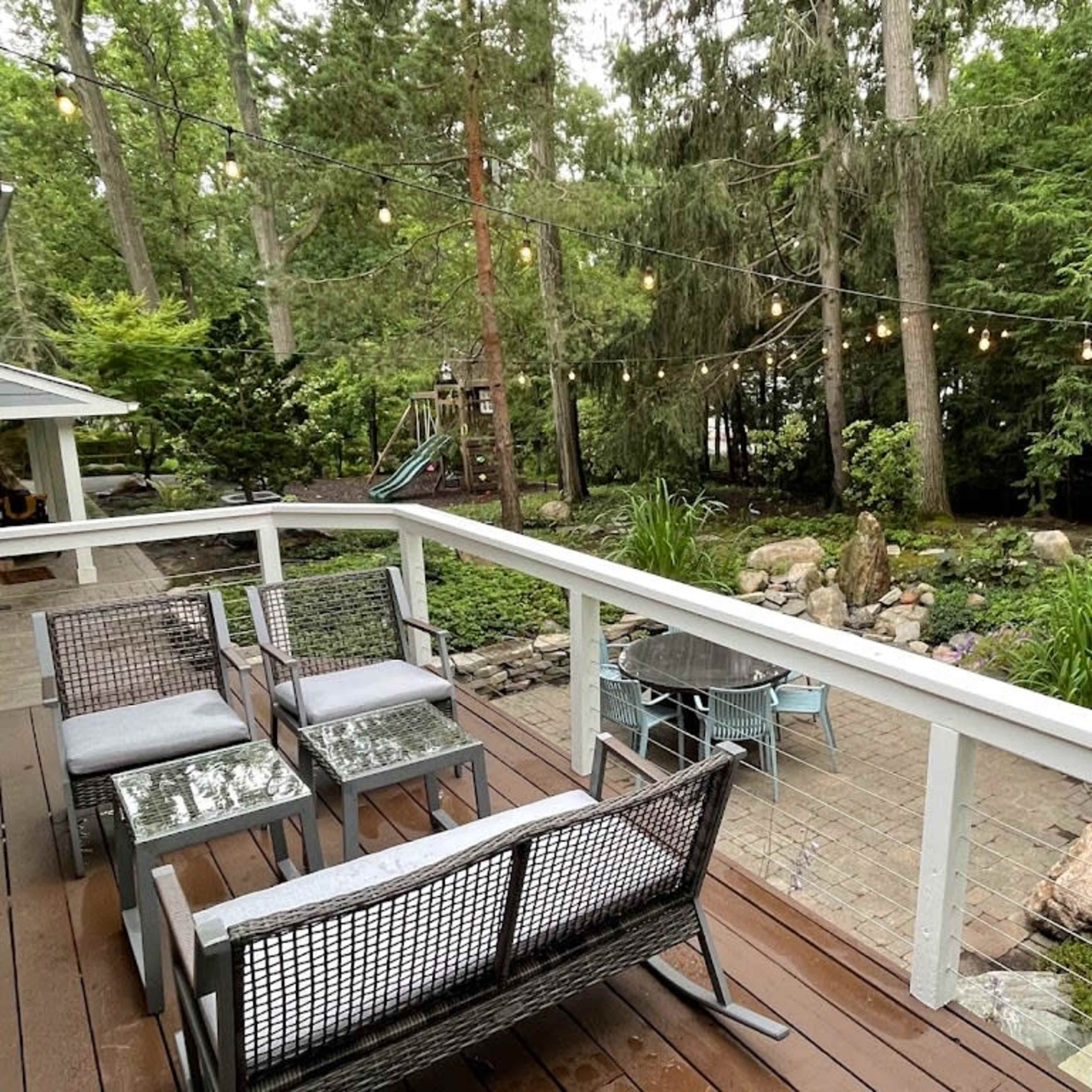 A wooden deck with seating overlooks a landscaped yard featuring trees, rocks, and garden lights.