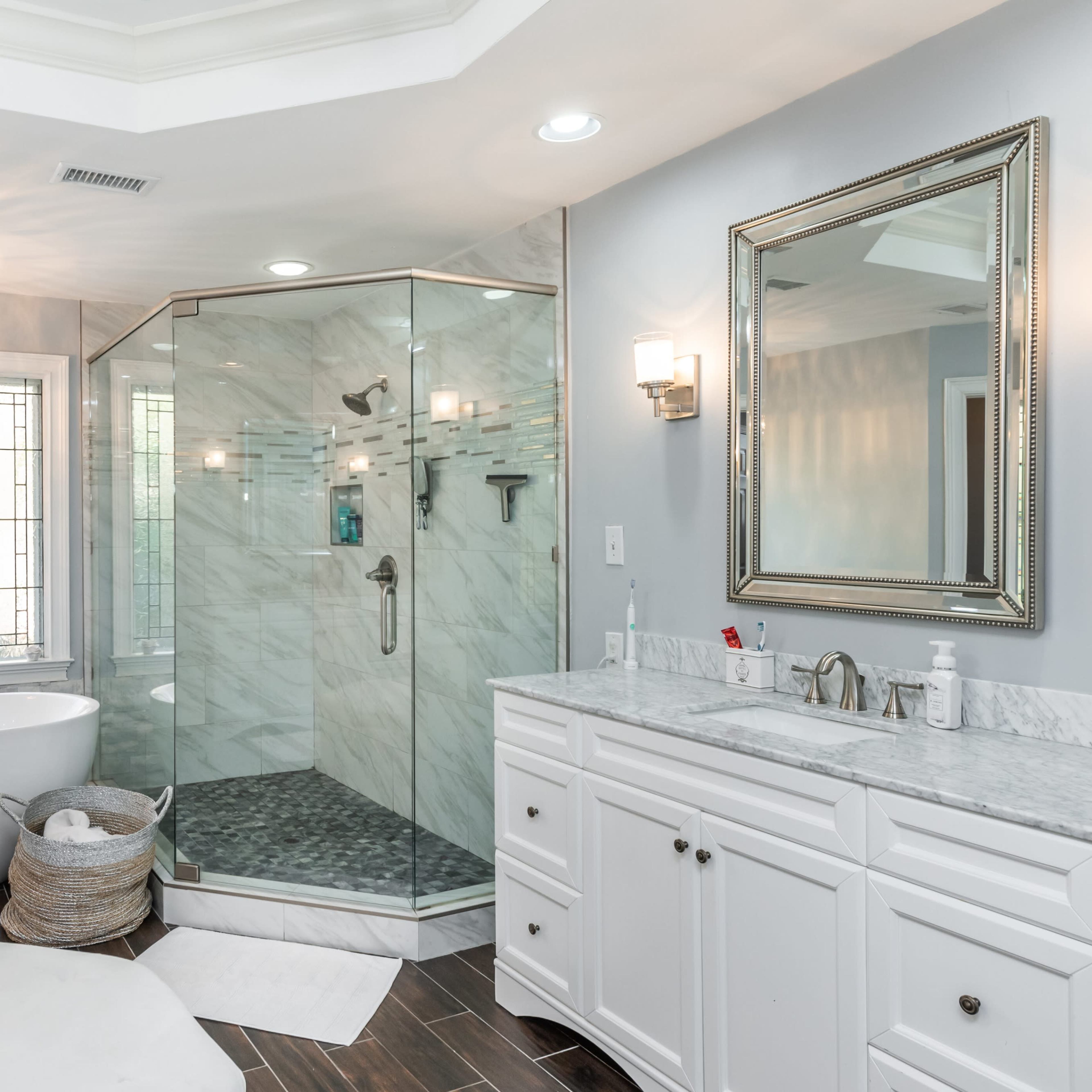 A modern bathroom featuring a freestanding bathtub, a glass shower enclosure, a double vanity with marble countertops, and a decorative window.
