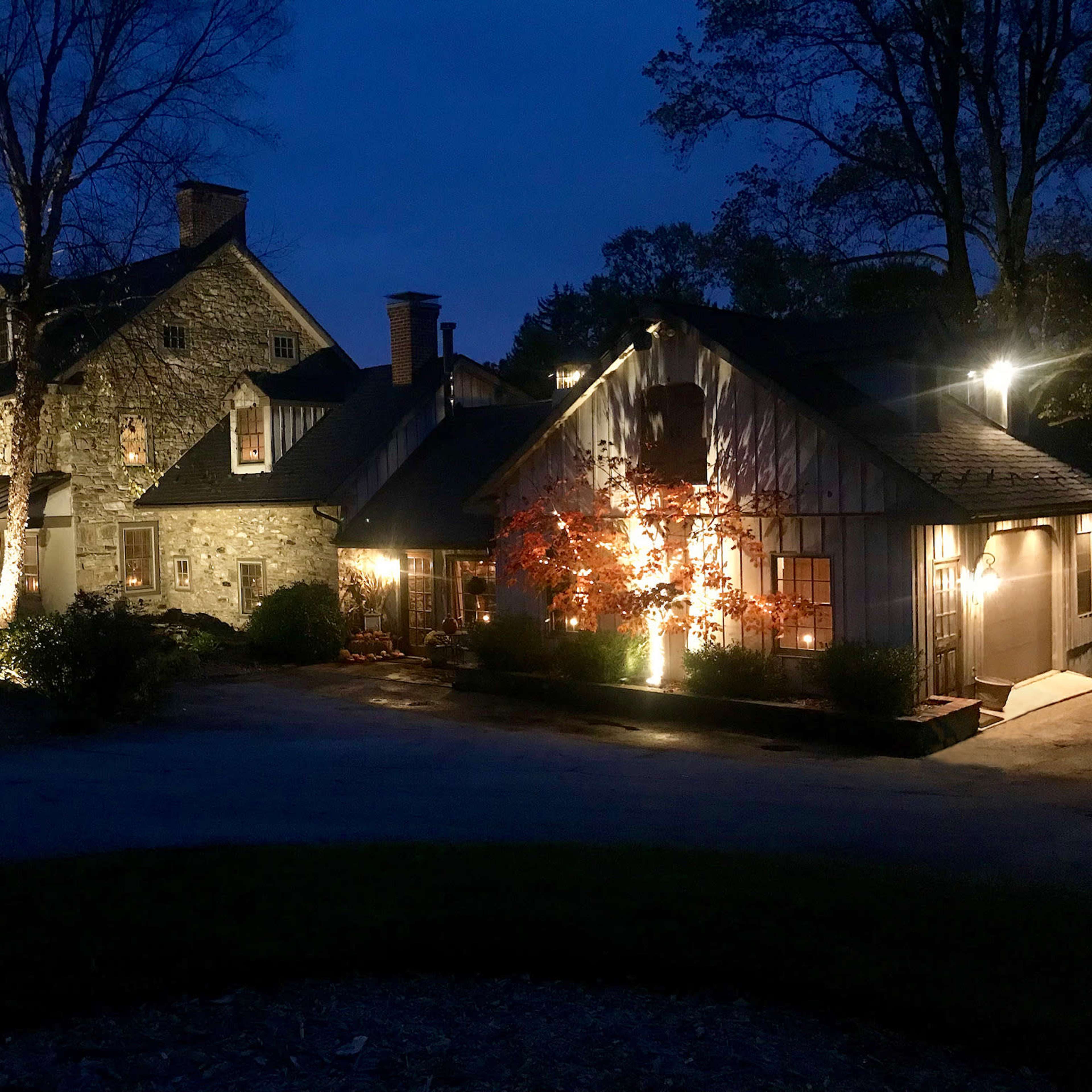 A stone and wood house is illuminated by lights at dusk, with trees and shrubs in the foreground.