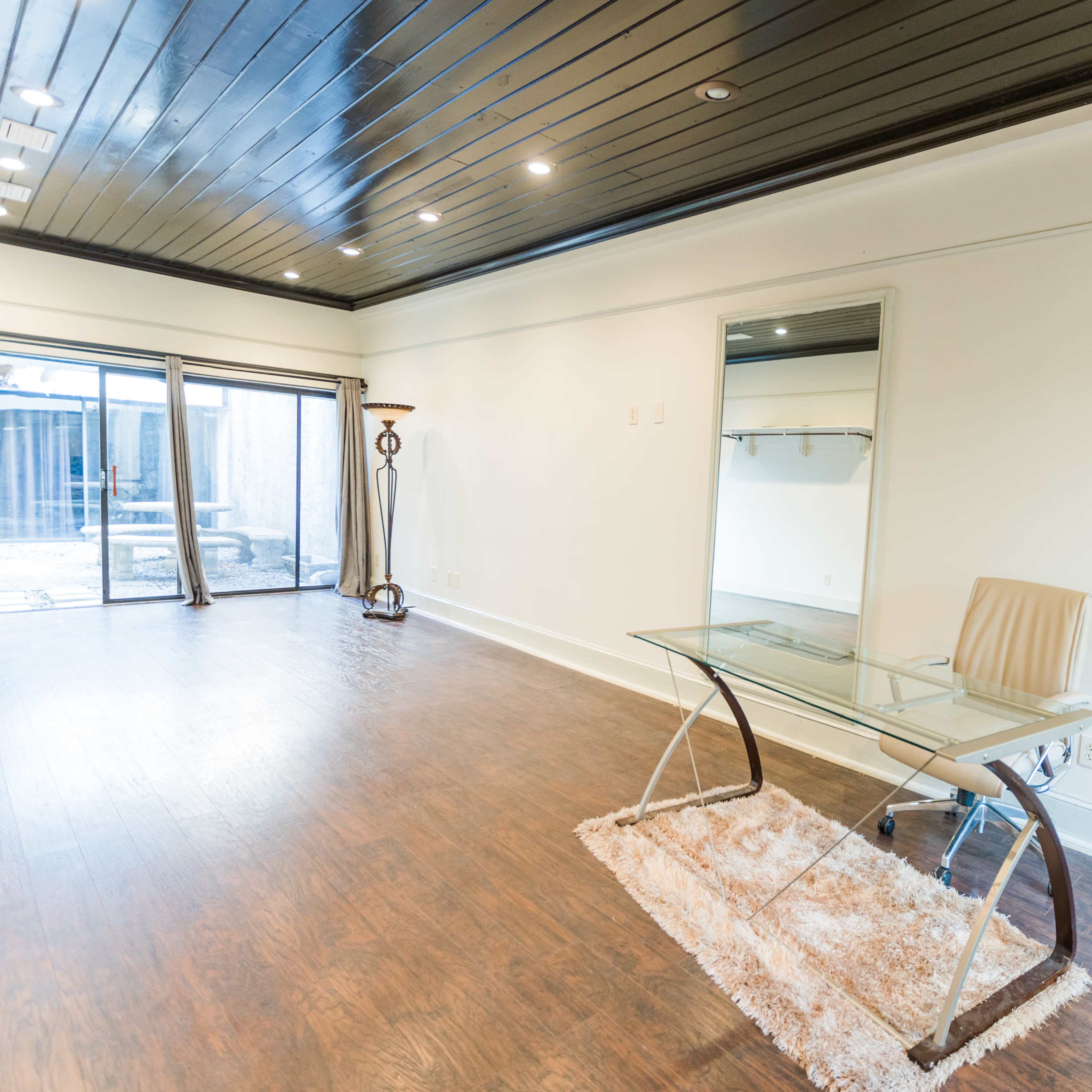 A spacious room with a glass-top desk and a chair on a rug, featuring large windows that lead to an outdoor area.