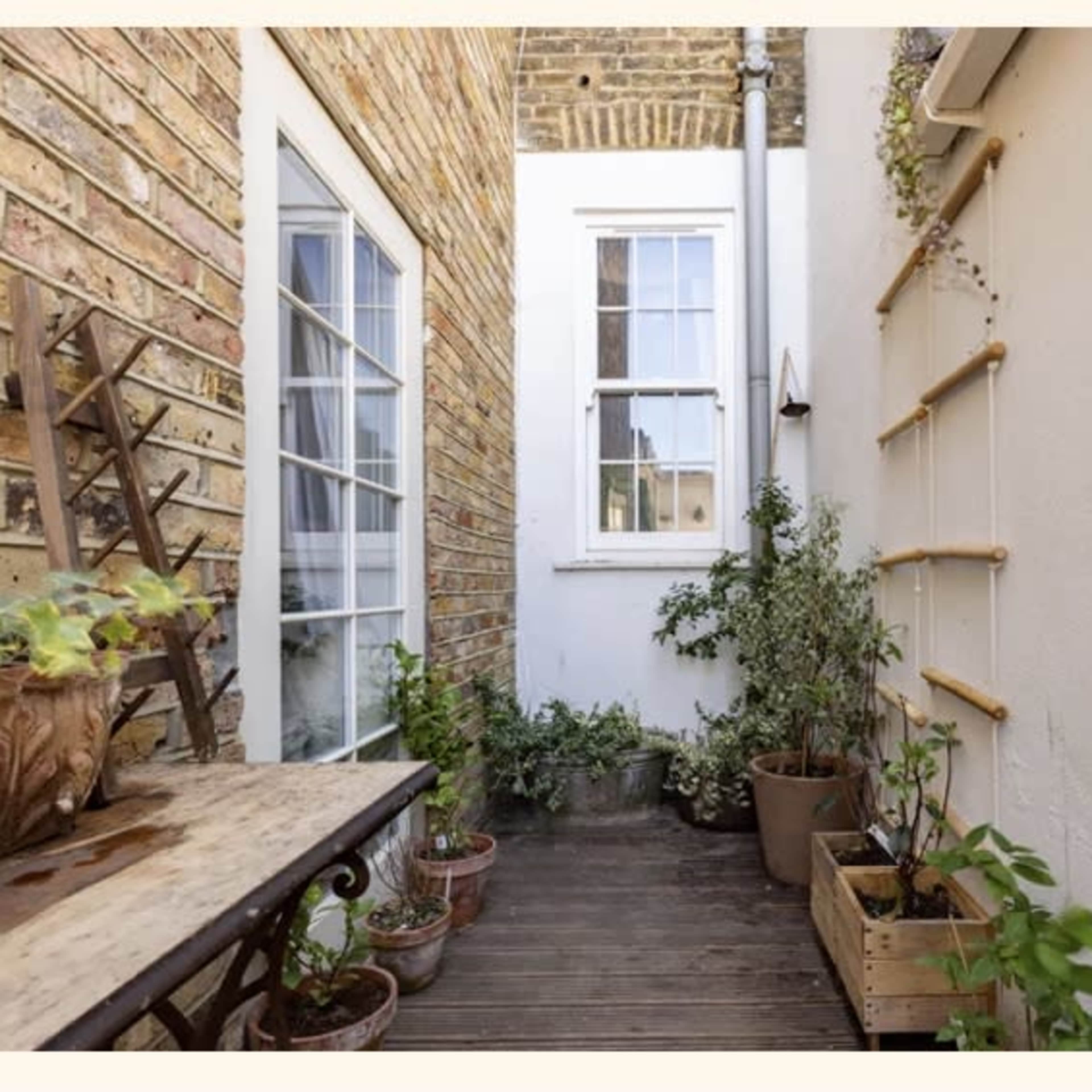 A narrow outdoor space features potted plants, a wooden table, and brick walls.