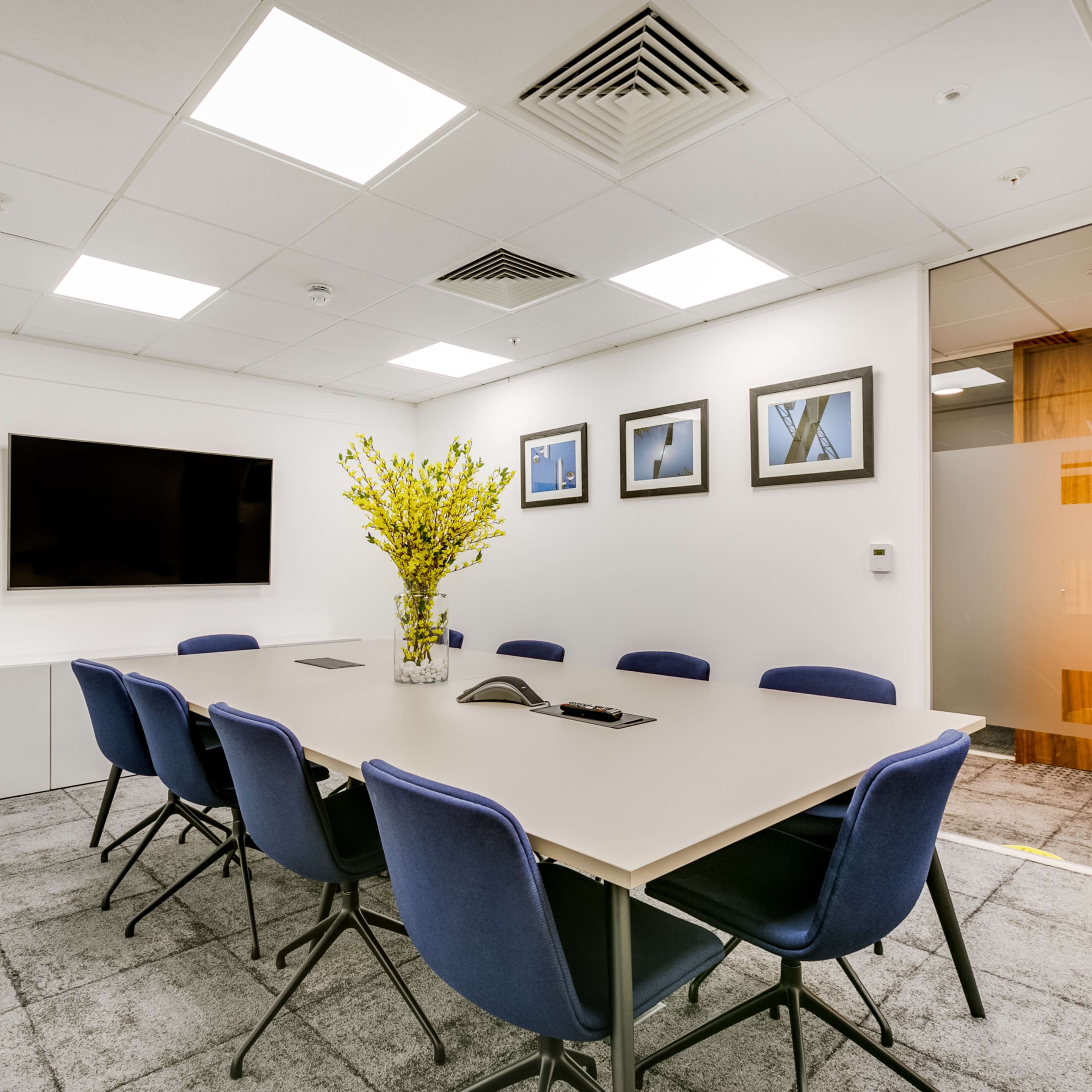 A modern conference room features a long table surrounded by blue chairs, a television on the wall, and a large flower arrangement on the table.