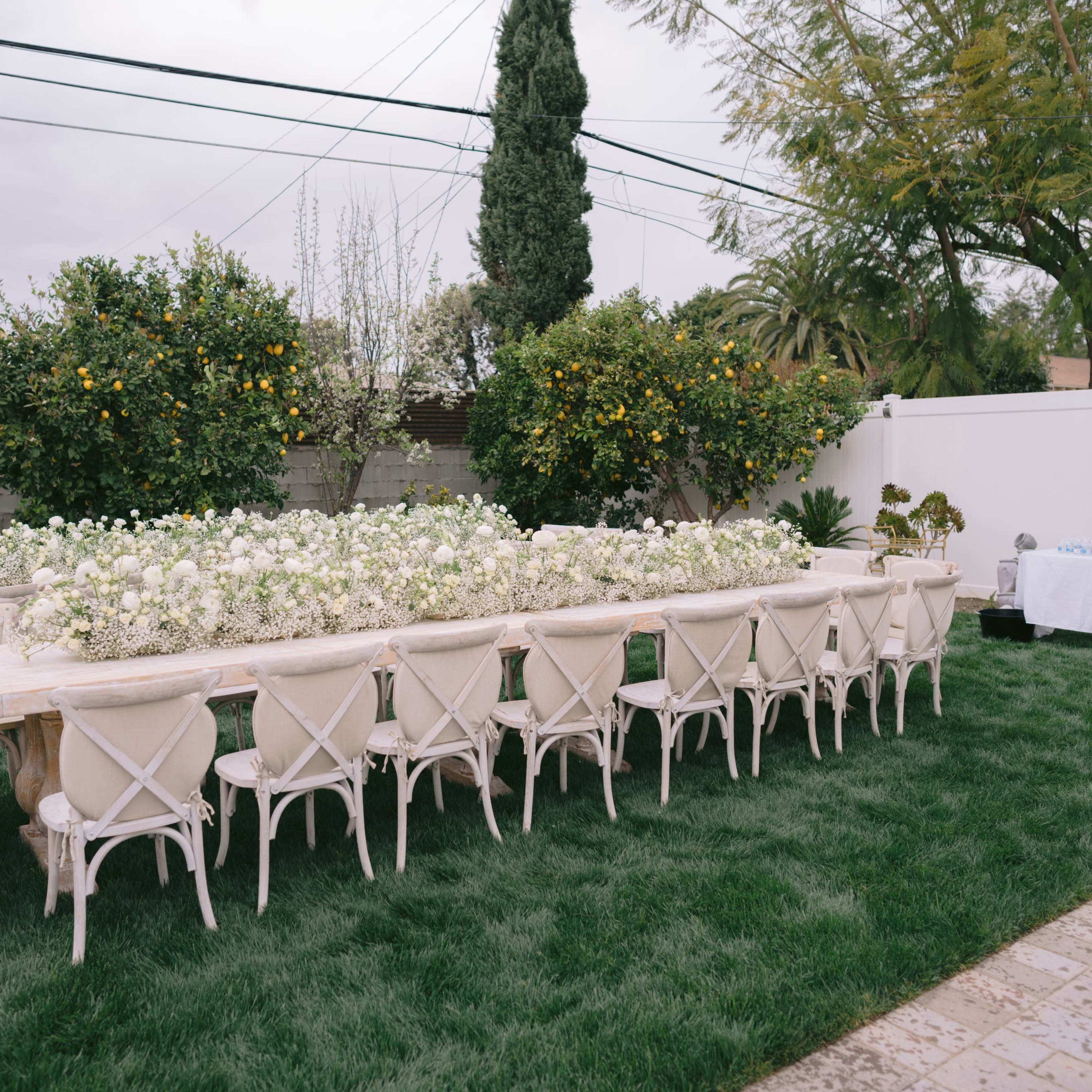 A long outdoor table decorated with white flowers is set up on a grassy area, surrounded by trees and a decorative table in the background.