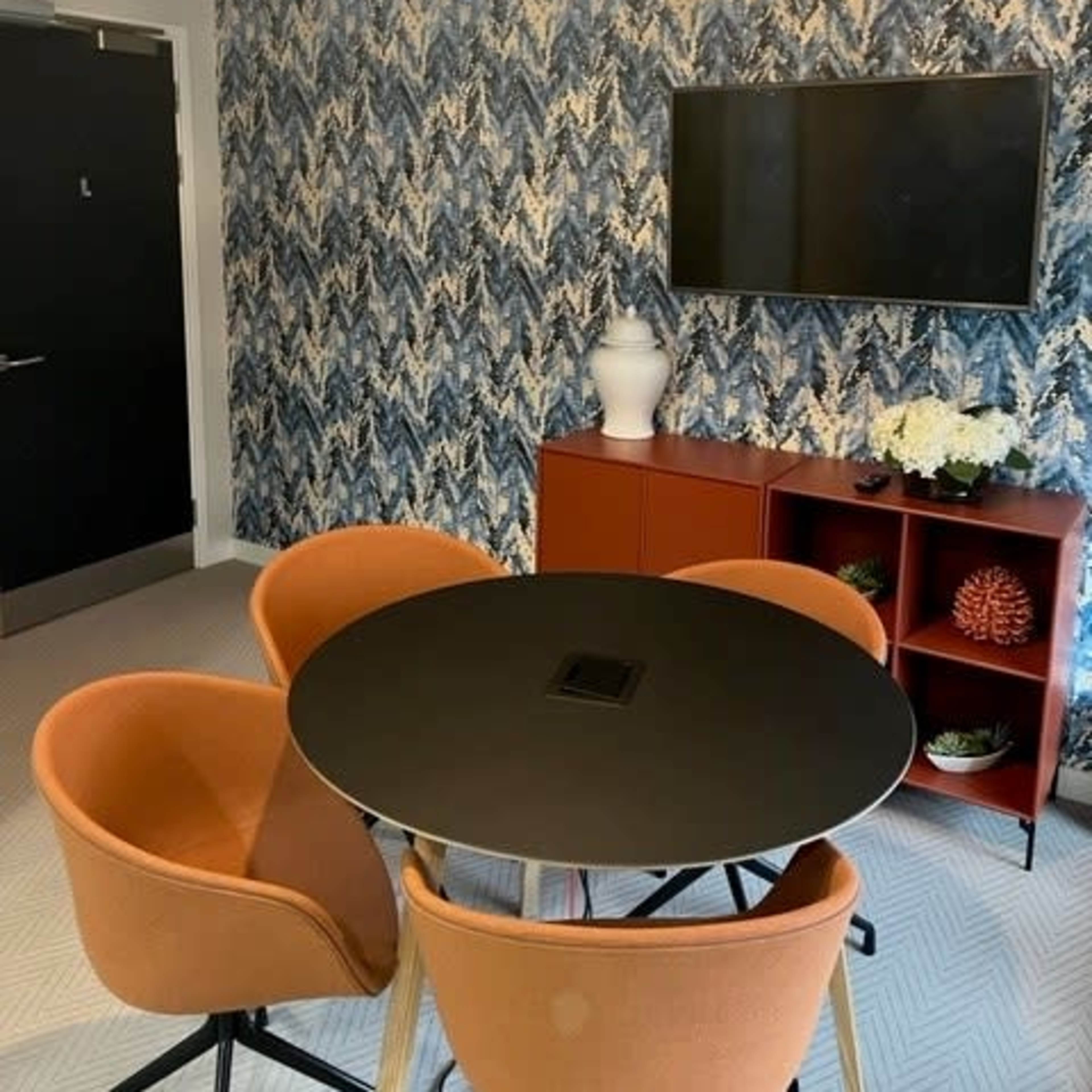A modern meeting room features a round black table surrounded by orange chairs, a patterned blue wallpaper, and a television mounted on the wall.