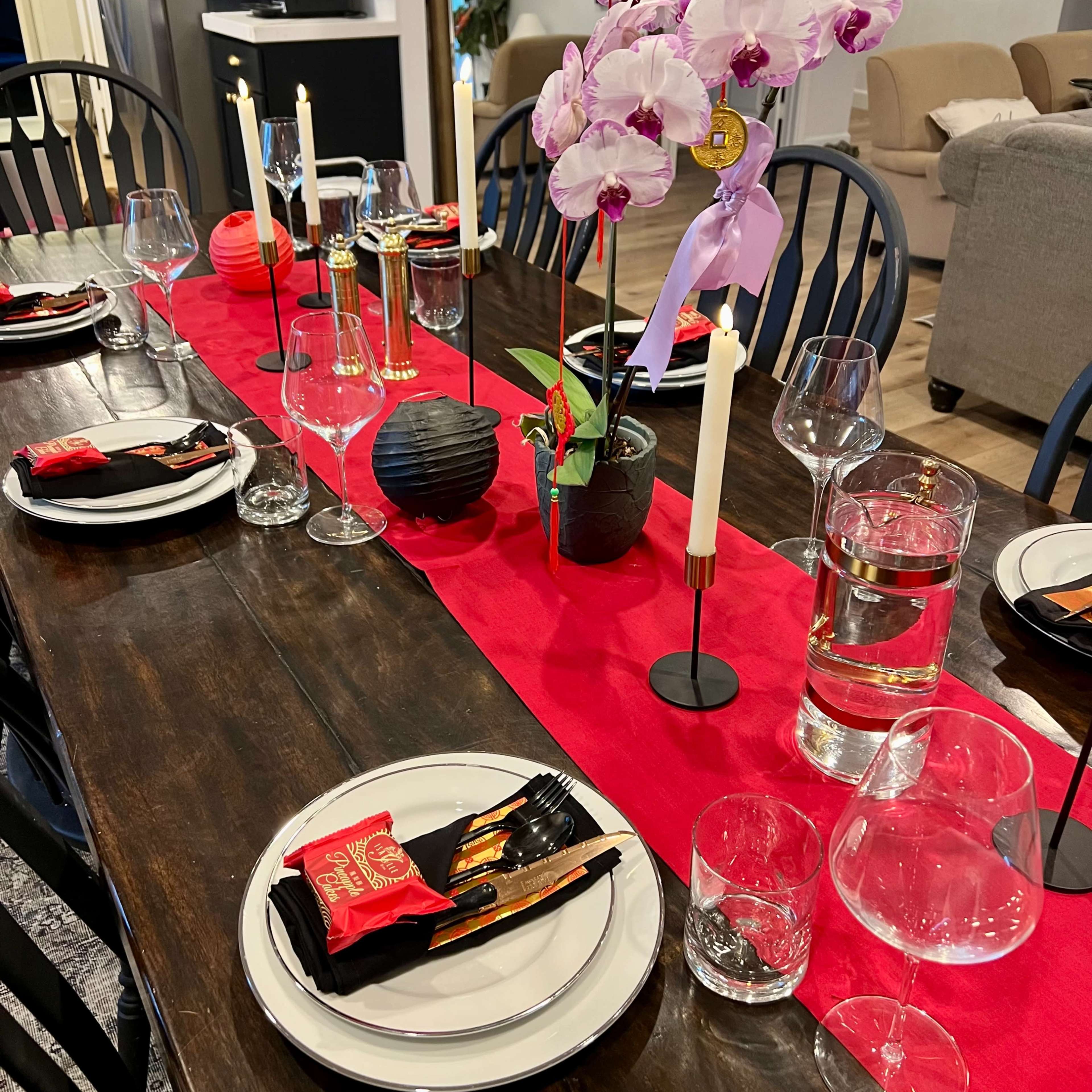 A dining table is set with plates, glasses, and silverware, featuring a red table runner adorned with an orchid centerpiece and candles.