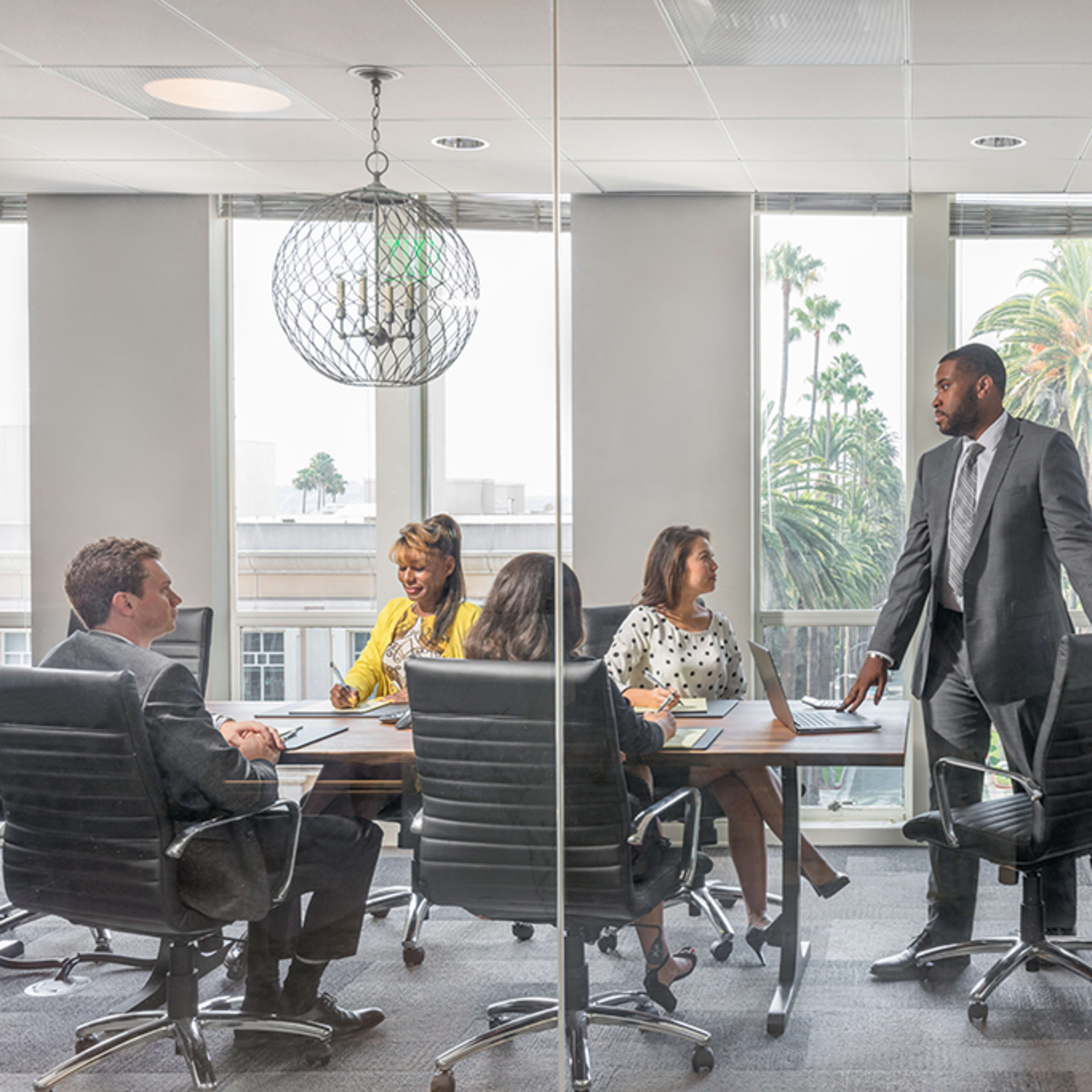 A group of six professionals is seated around a conference table in a modern office with large windows overlooking palm trees.