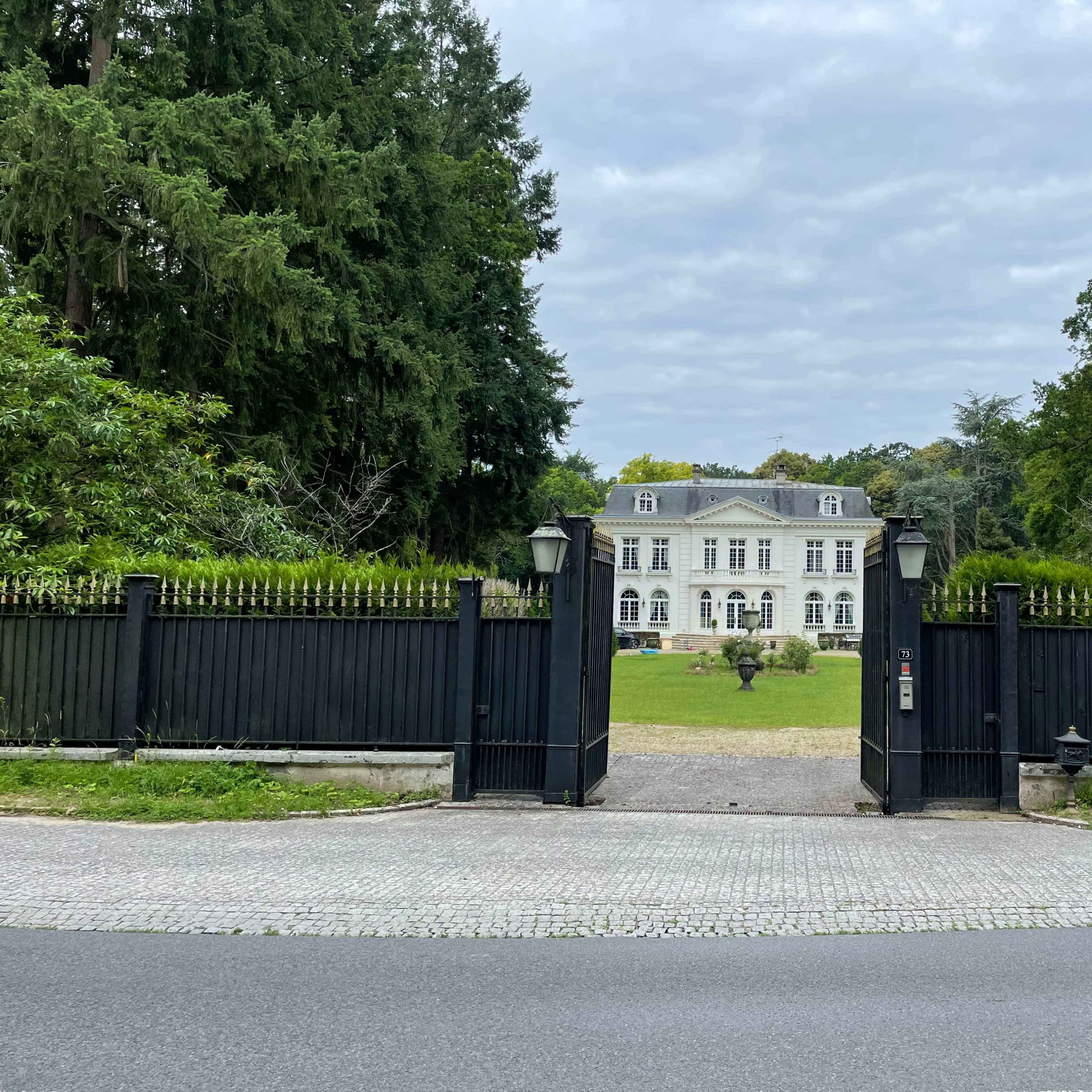 A large white mansion is visible through a black decorative gate, surrounded by green trees and a manicured lawn.