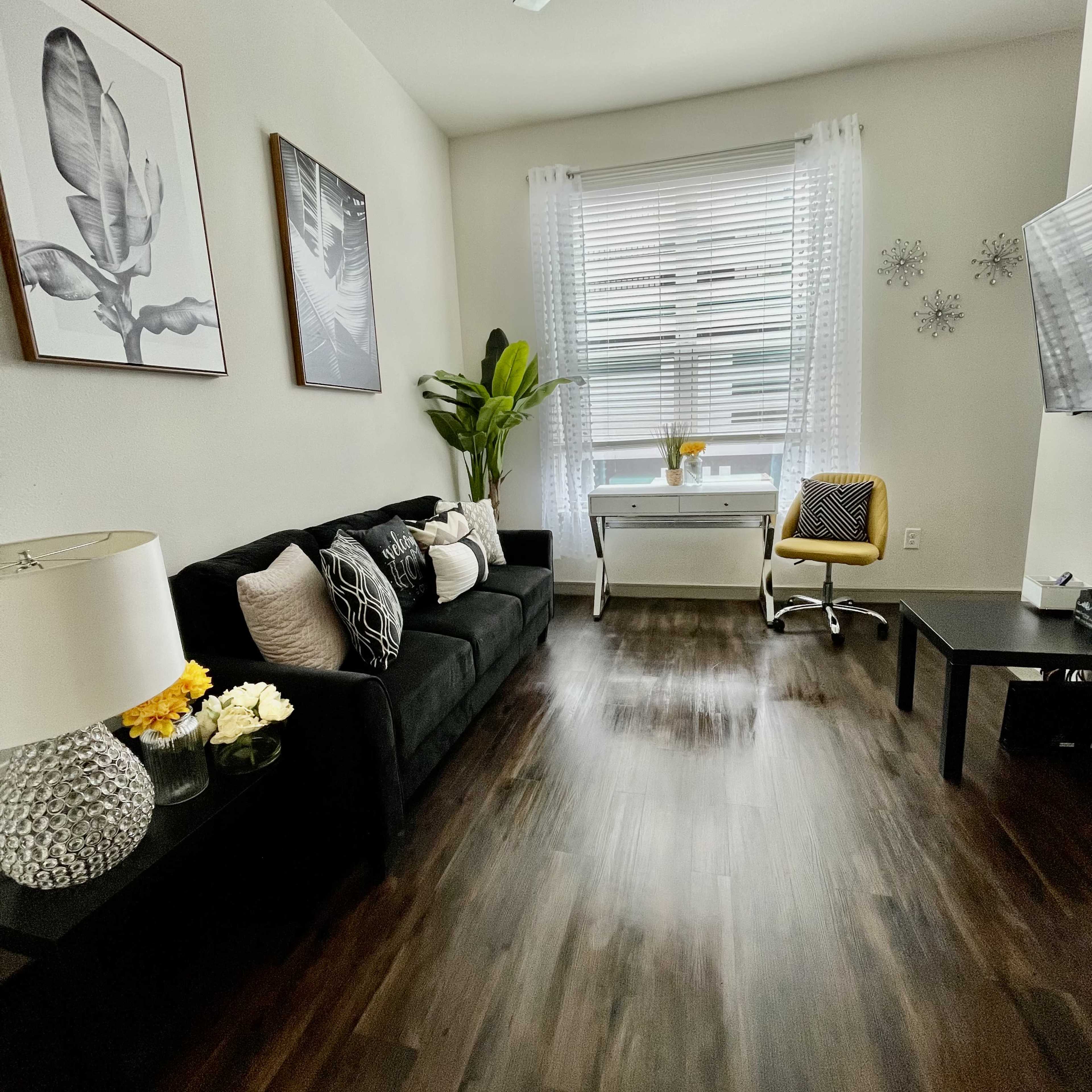 The room features a black sofa, a small desk with a chair, and artwork on the walls, complemented by a large window with sheer curtains.