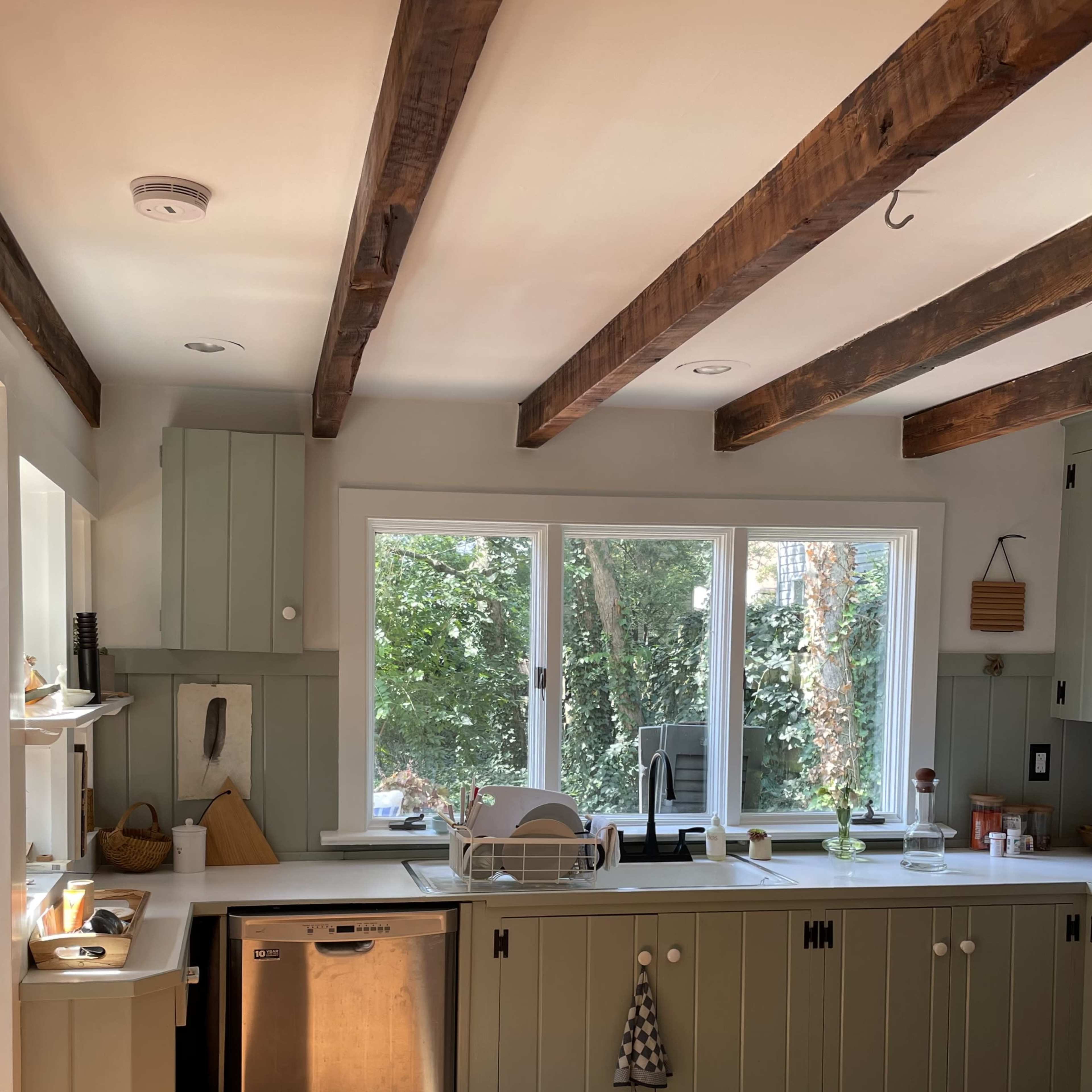 A kitchen features green cabinetry, wooden beams, and a window overlooking a garden.