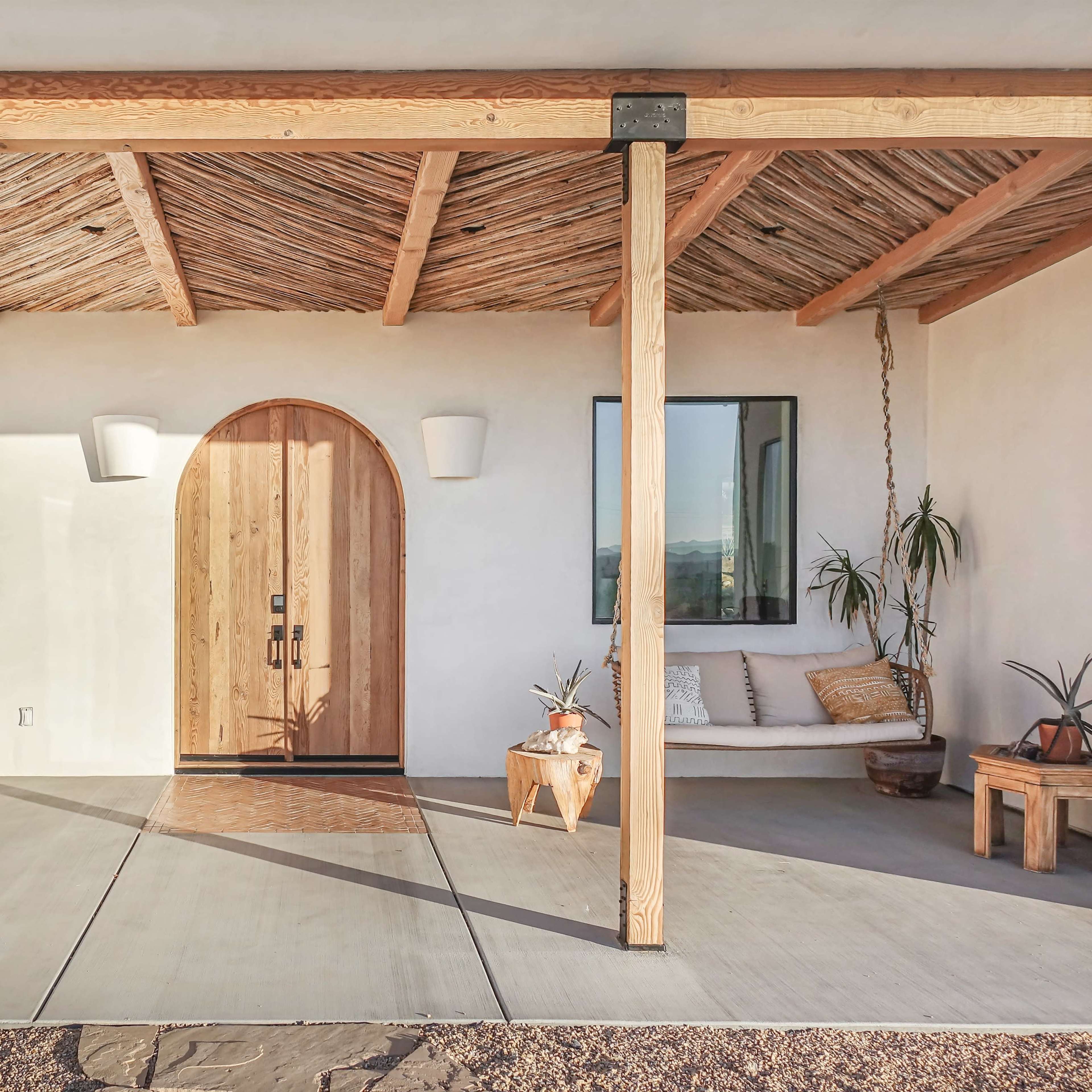 A modern entrance with a wooden door framed by large, open beams, a thatched roof, and minimalist seating arrangements on a concrete patio.