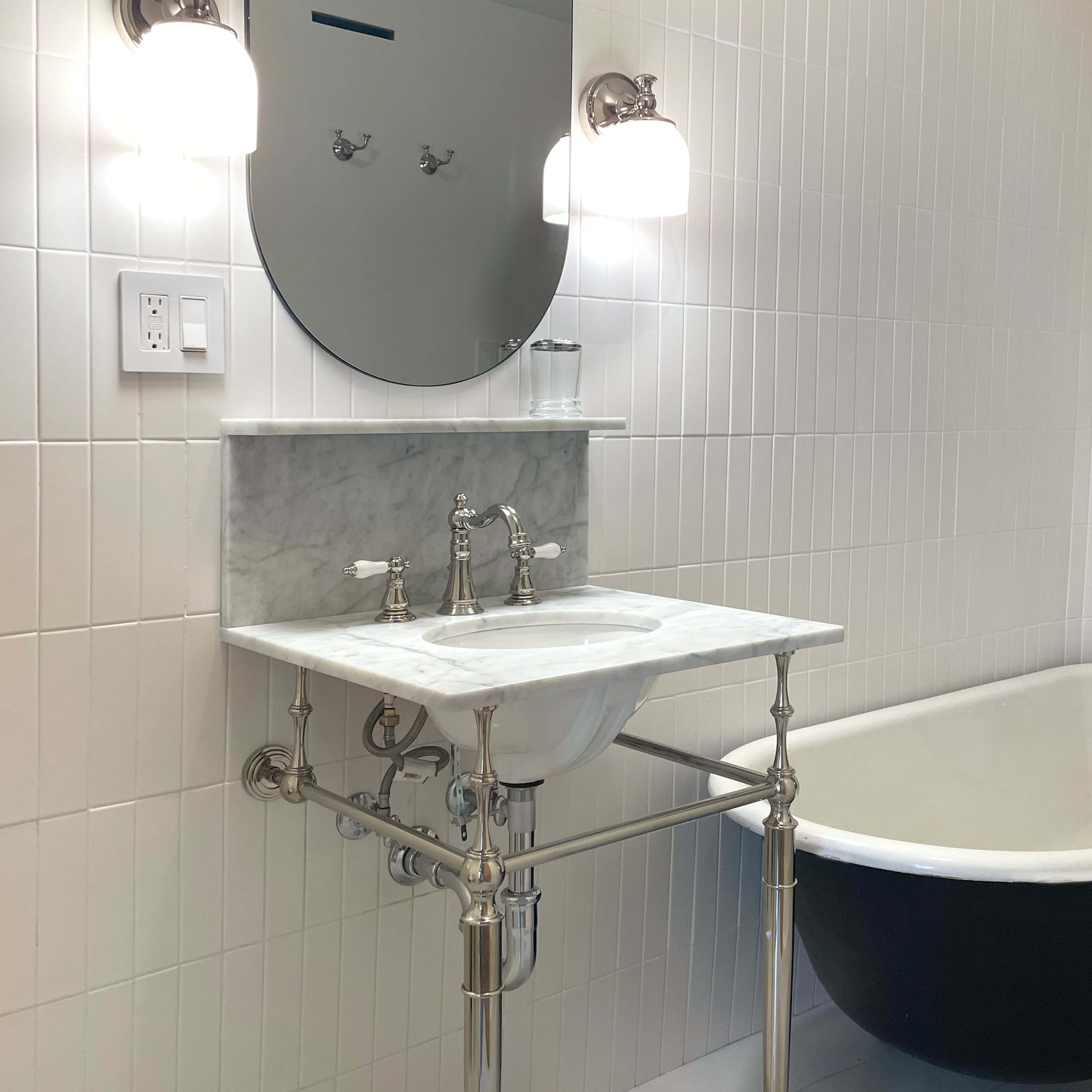 A marble sink with chrome fixtures is mounted on a white tiled wall, accompanied by a large round mirror and a vintage-style freestanding bathtub.