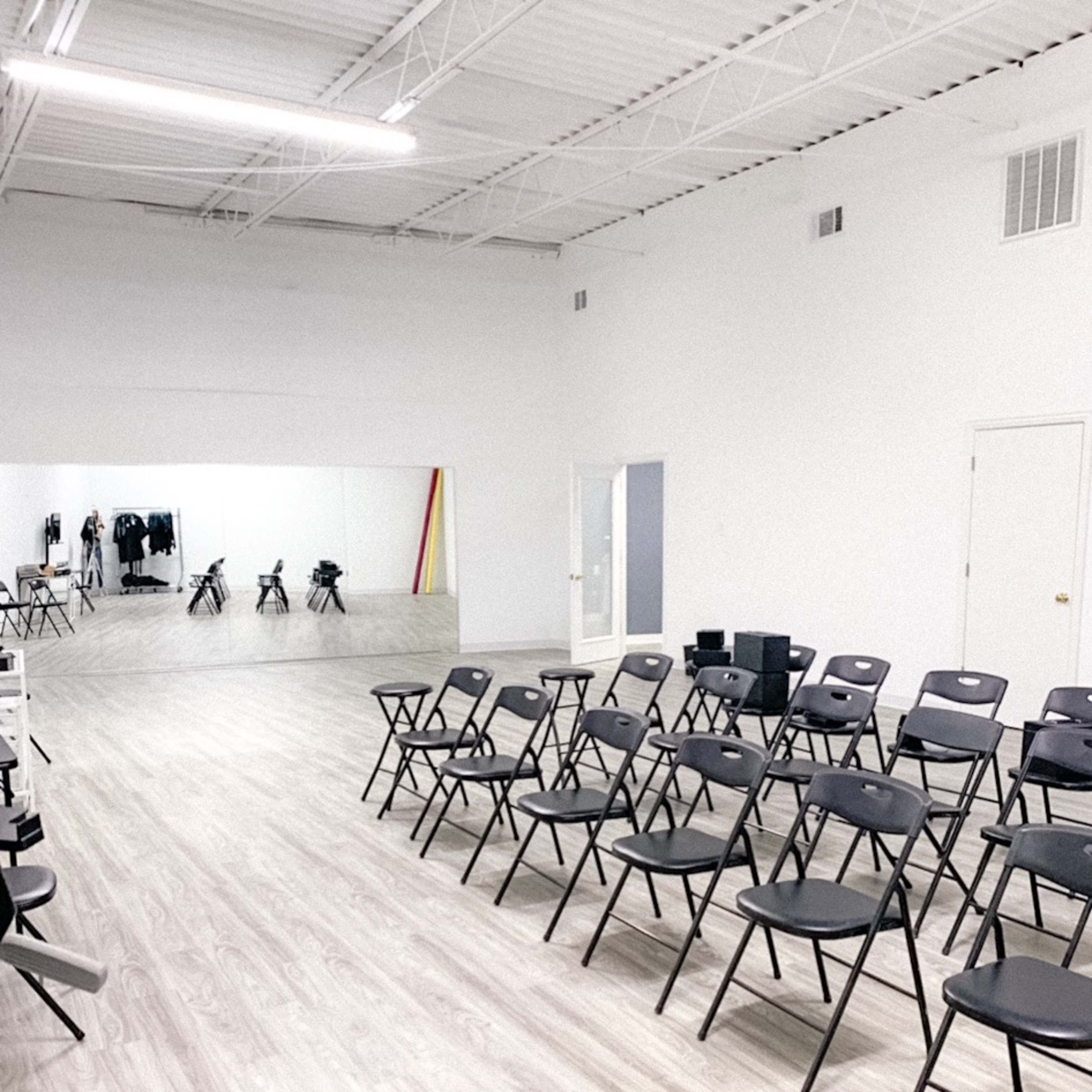 A spacious room with rows of black folding chairs facing a large mirror on a white wall.