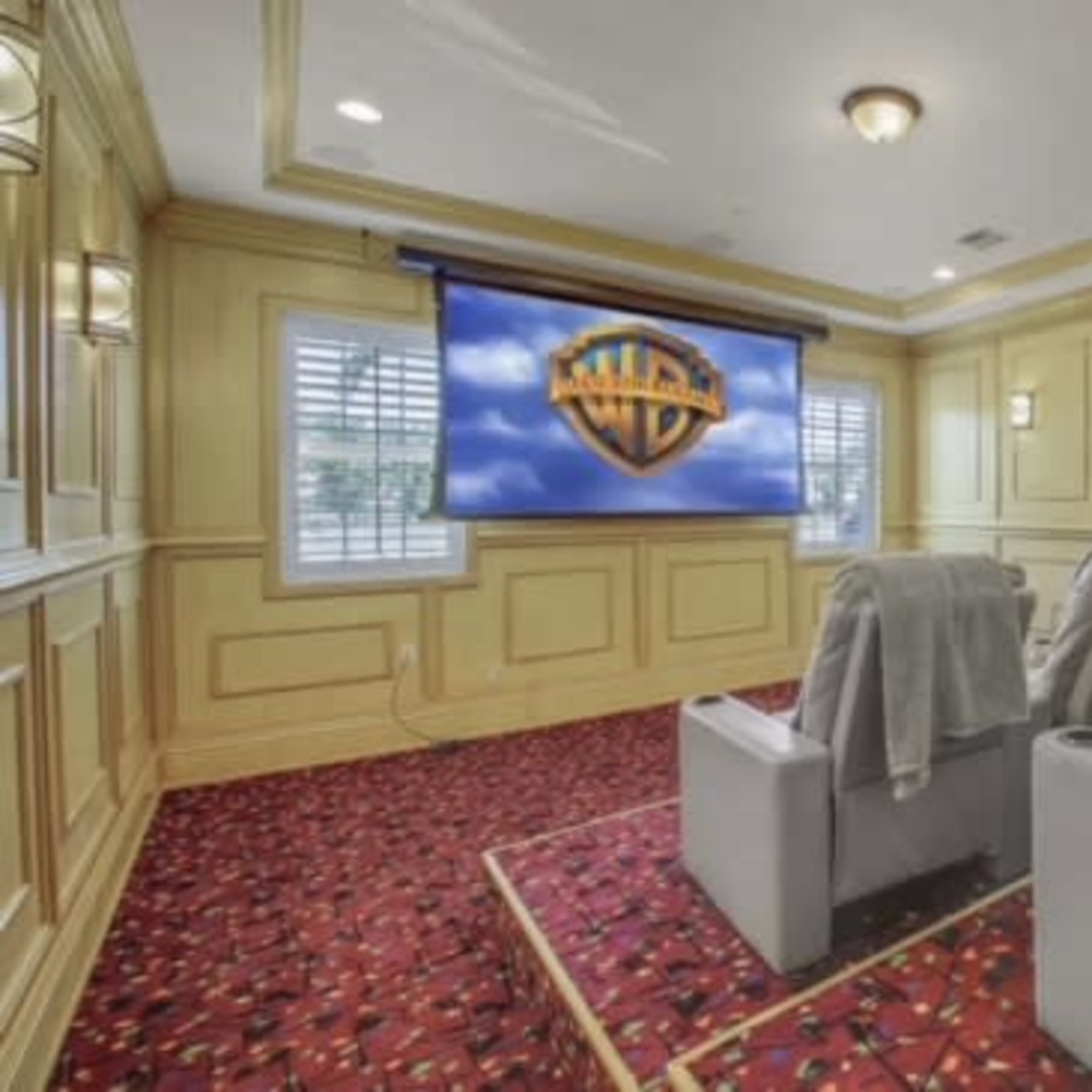 A home theater room features three reclining chairs with blankets facing a large screen displaying the Warner Bros. logo.