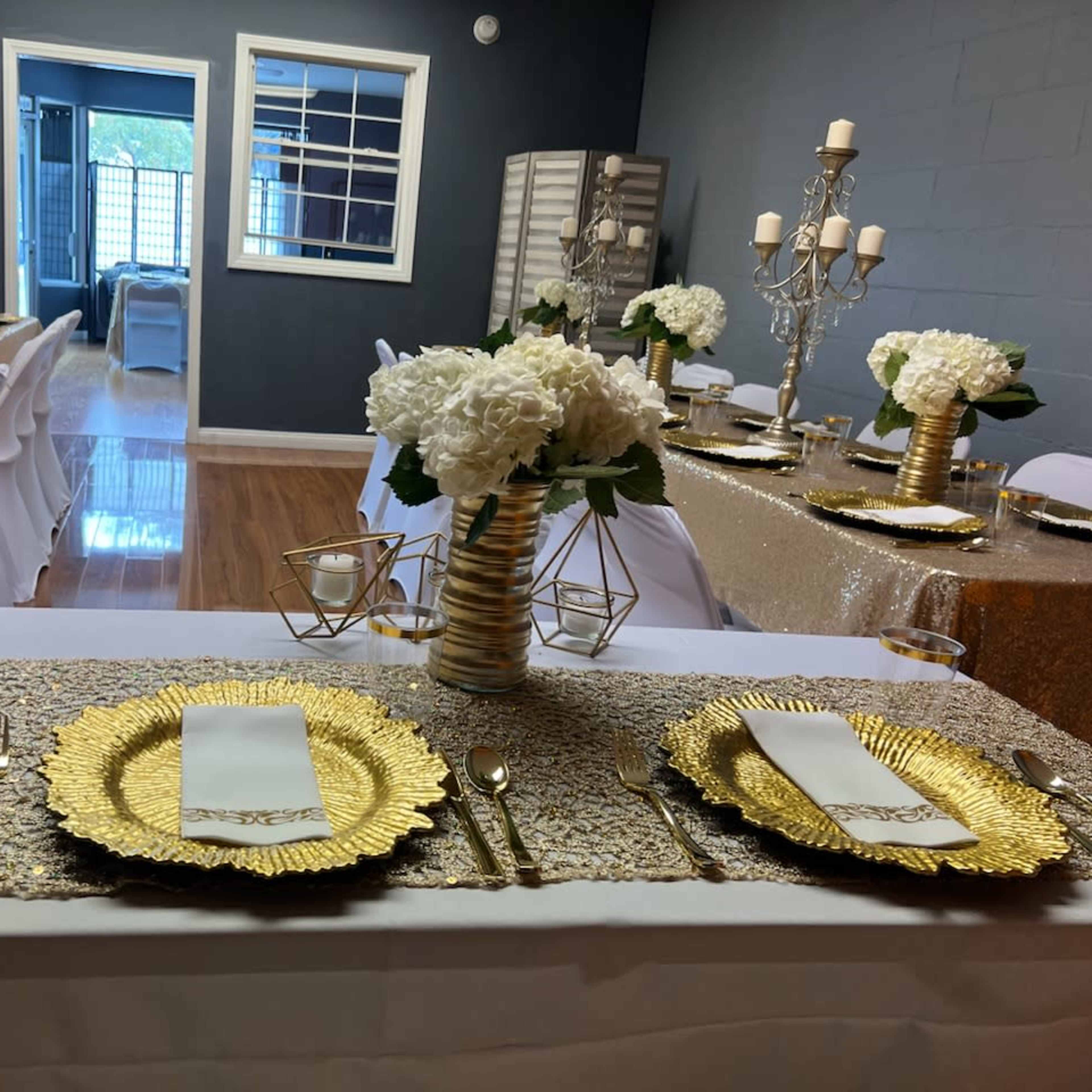 A decorated dining area features tables set with gold tableware, white floral centerpieces, and elegant candle holders.
