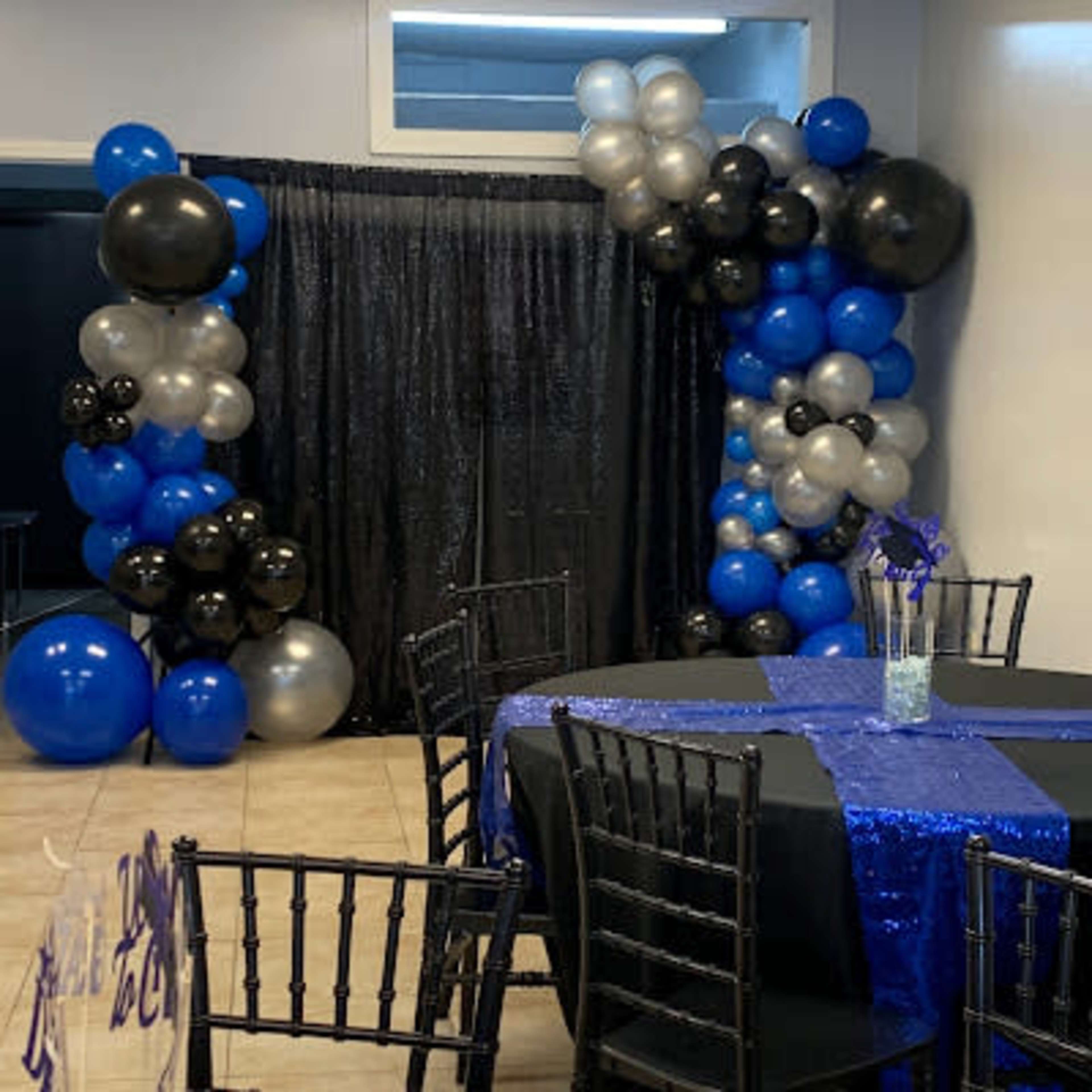 A party venue features a corner decorated with blue, black, and silver balloons beside a black curtain, with tables set in front covered in black and blue sequined tablecloths.