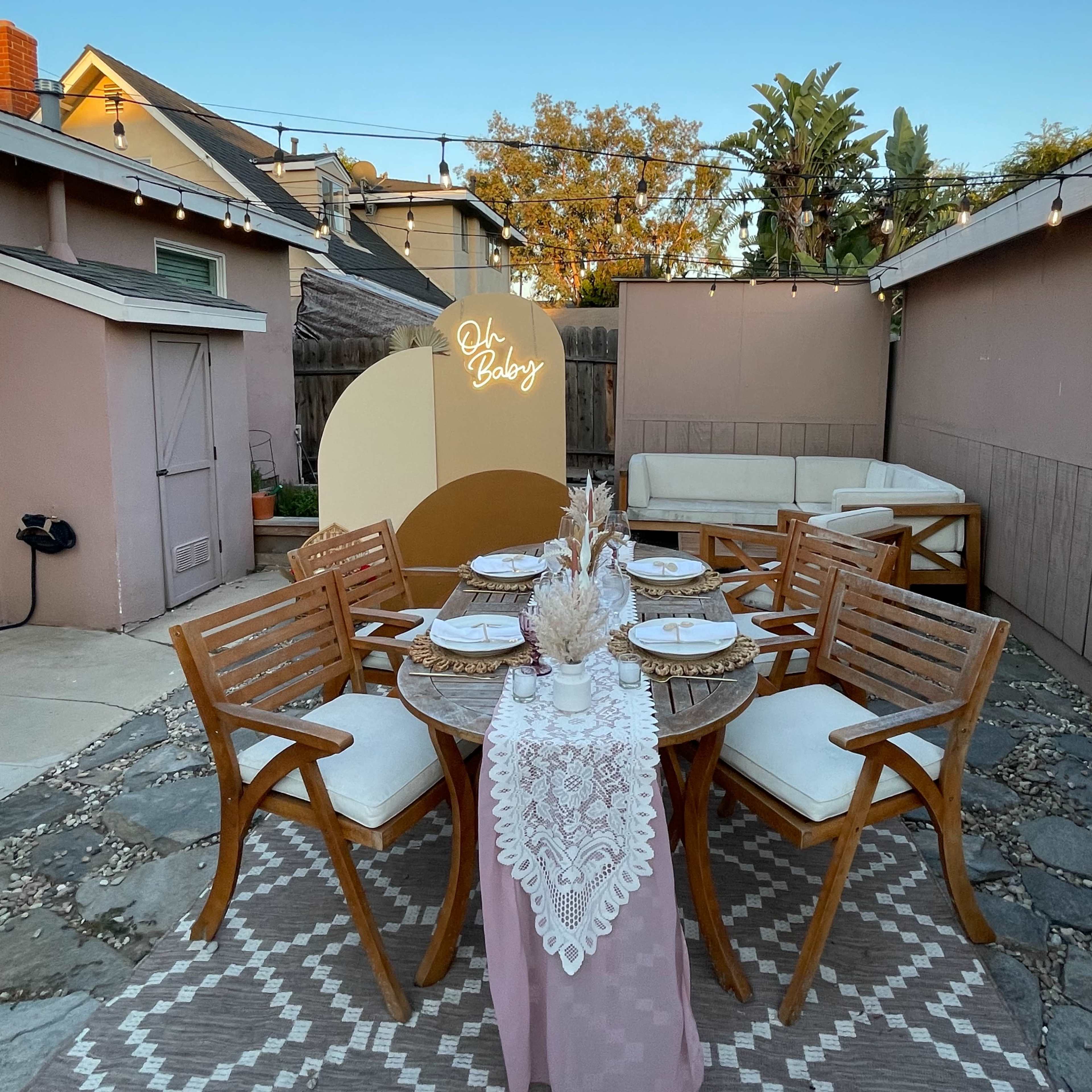 A stylish outdoor dining setup features a round table with lace decor, surrounded by wooden chairs, set against a backdrop of a cozy backyard with string lights and greenery.