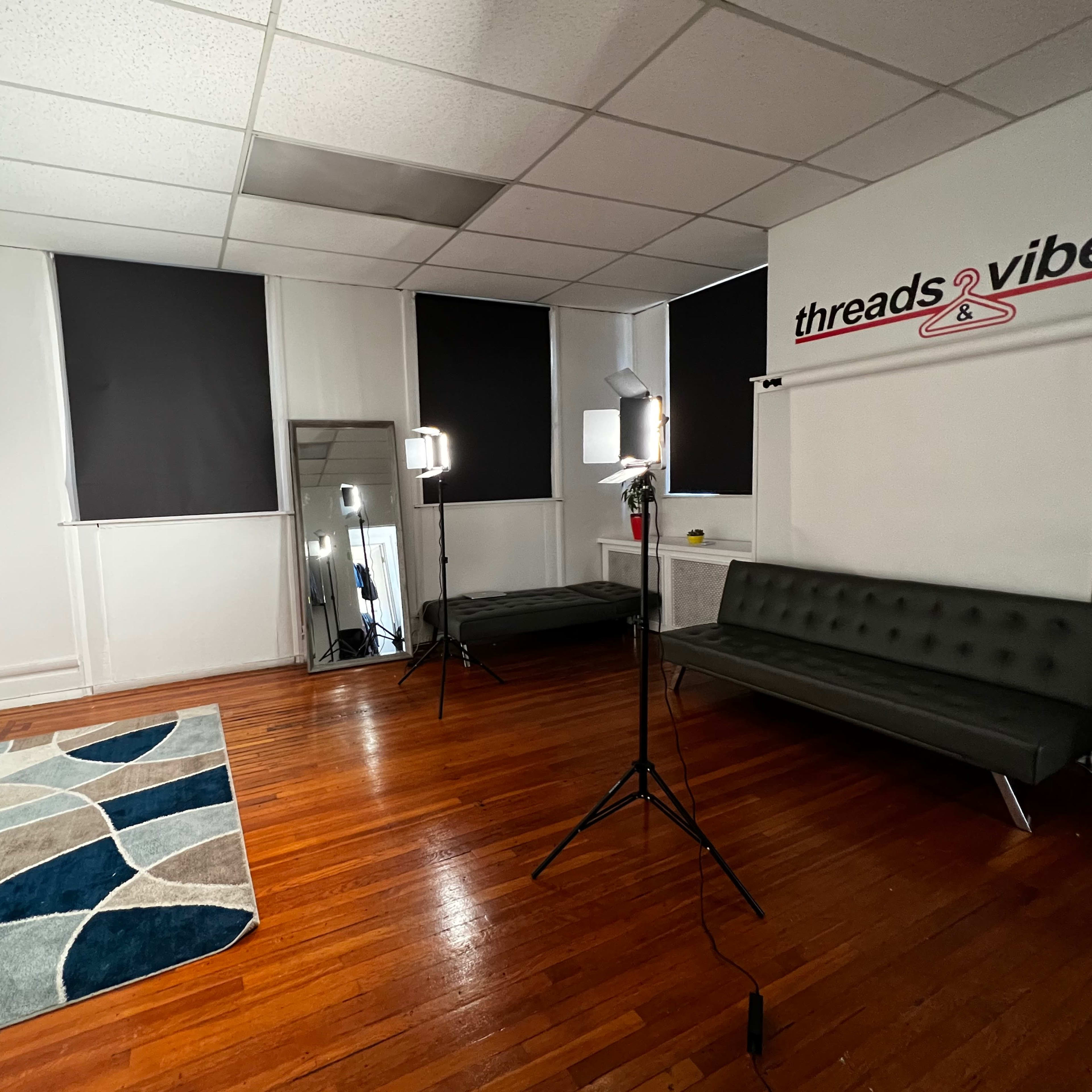 A well-lit studio space featuring a black sofa, a large mirror, and two light stands, with dark curtains covering the windows.