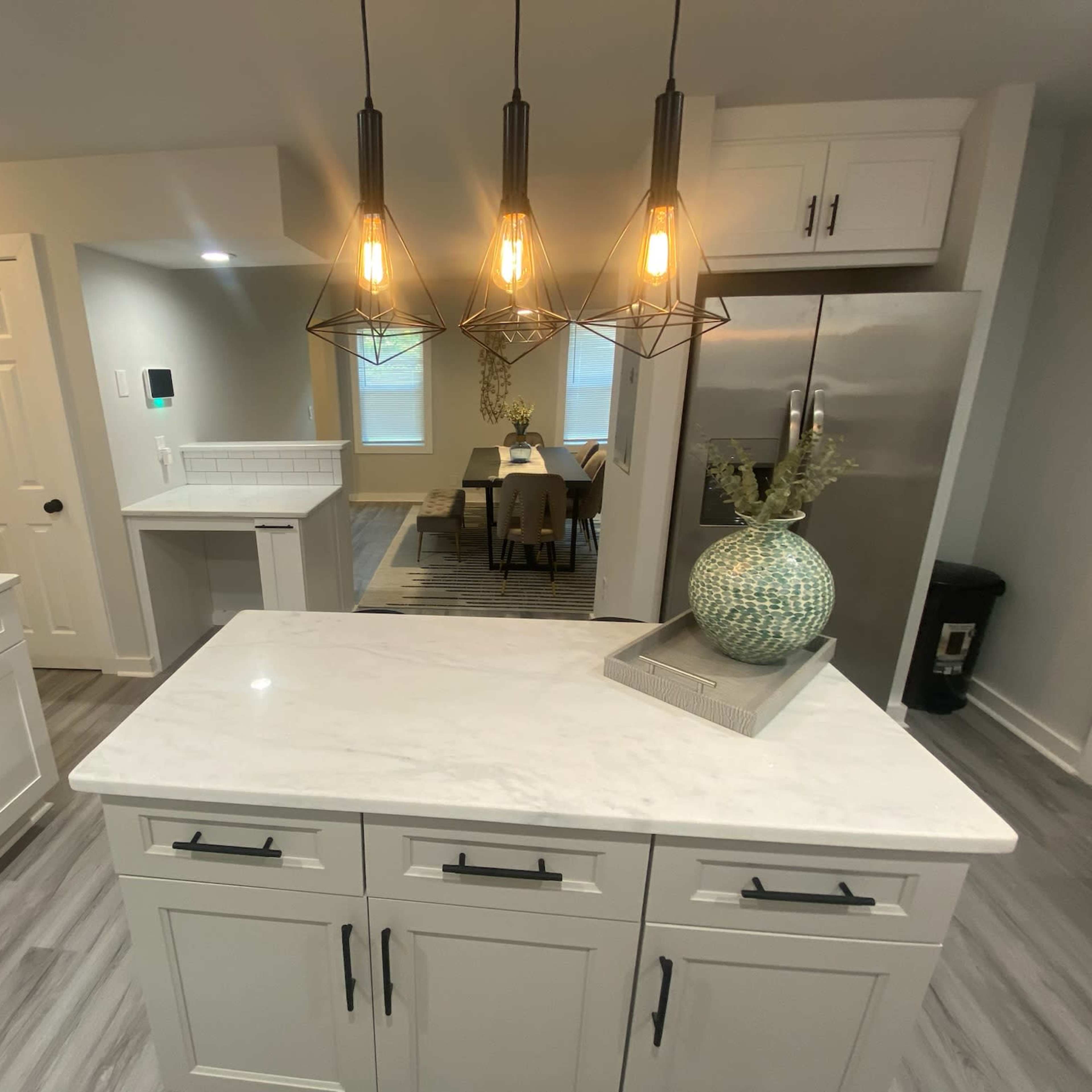 A modern kitchen with white cabinets, a marble island, pendant lighting, and a dining area visible in the background.