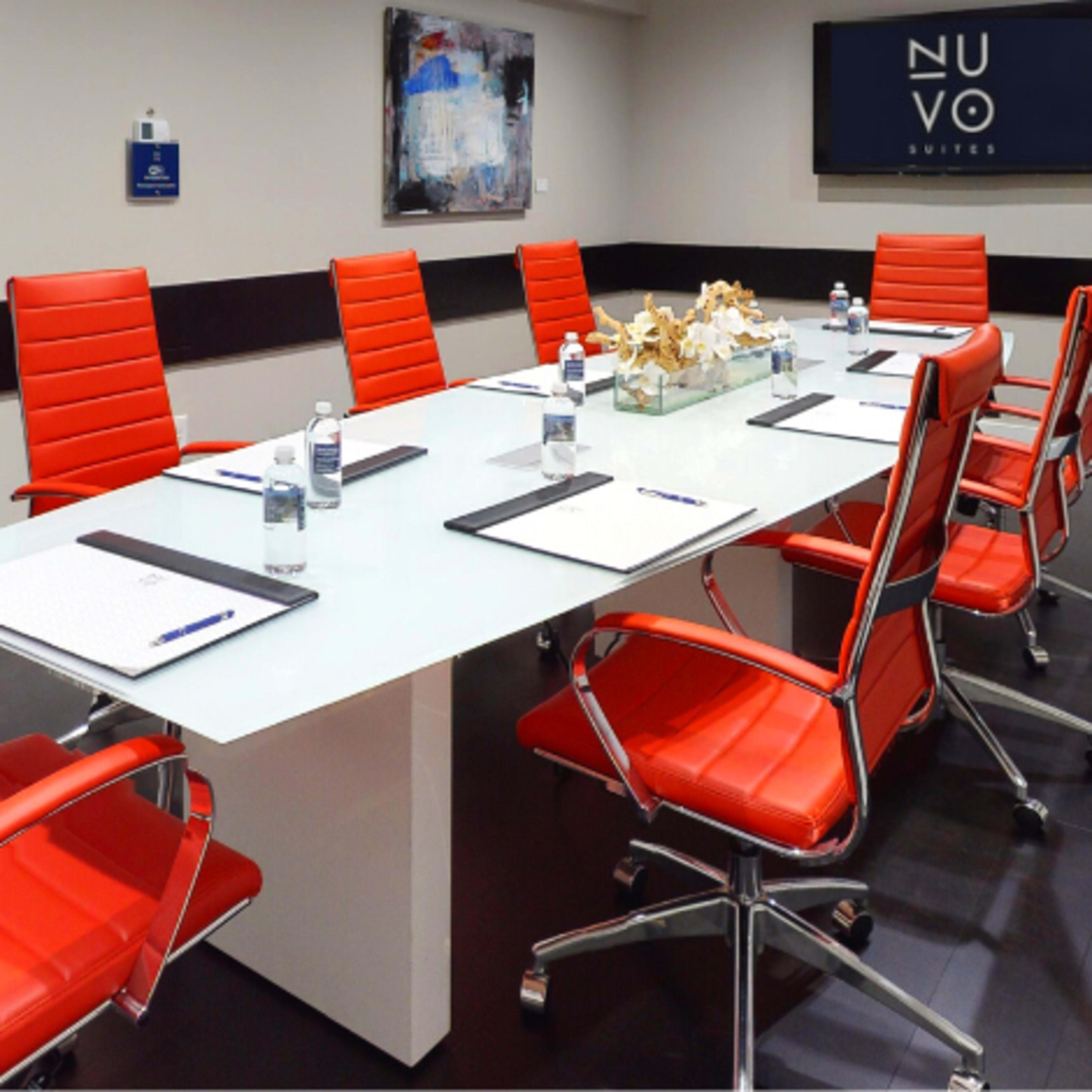 A modern conference room with a long glass table surrounded by red chairs, equipped with notepads and bottles of water.