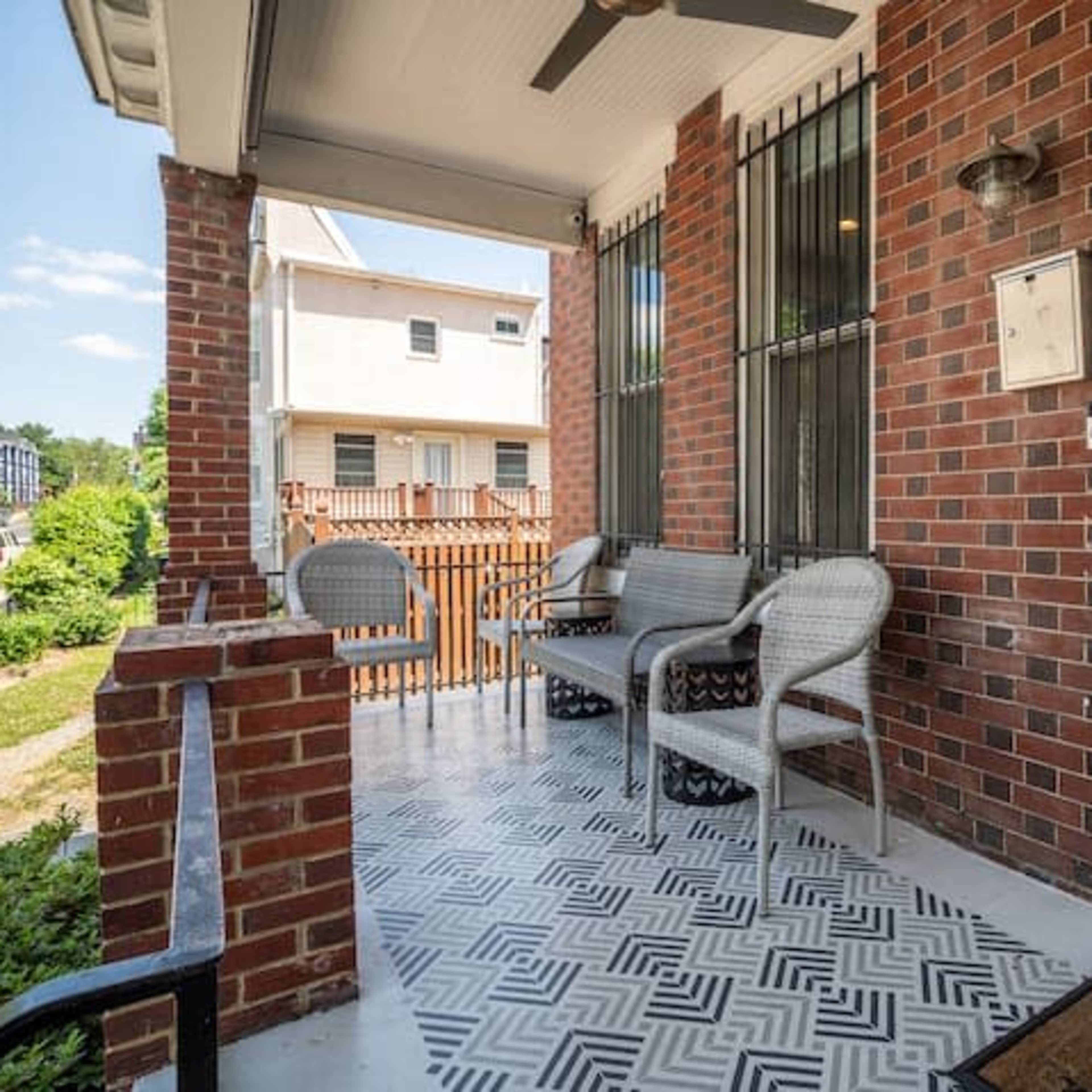 A brick front porch with a patterned tile floor, featuring a seating area of wicker chairs and a small table.