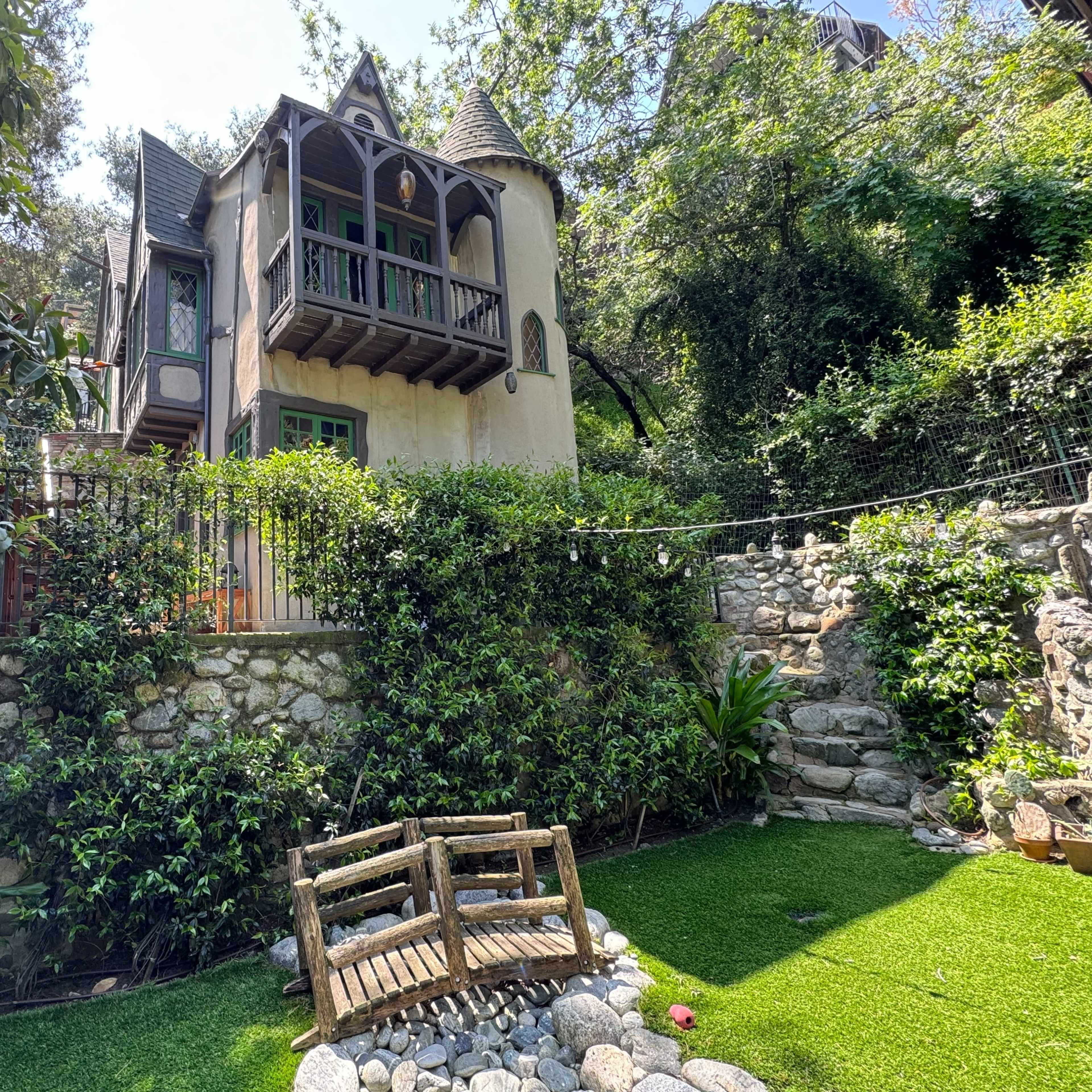 A two-story, Tudor-style house with a balcony is surrounded by lush greenery and a stone wall, featuring a small garden with a wooden bench.