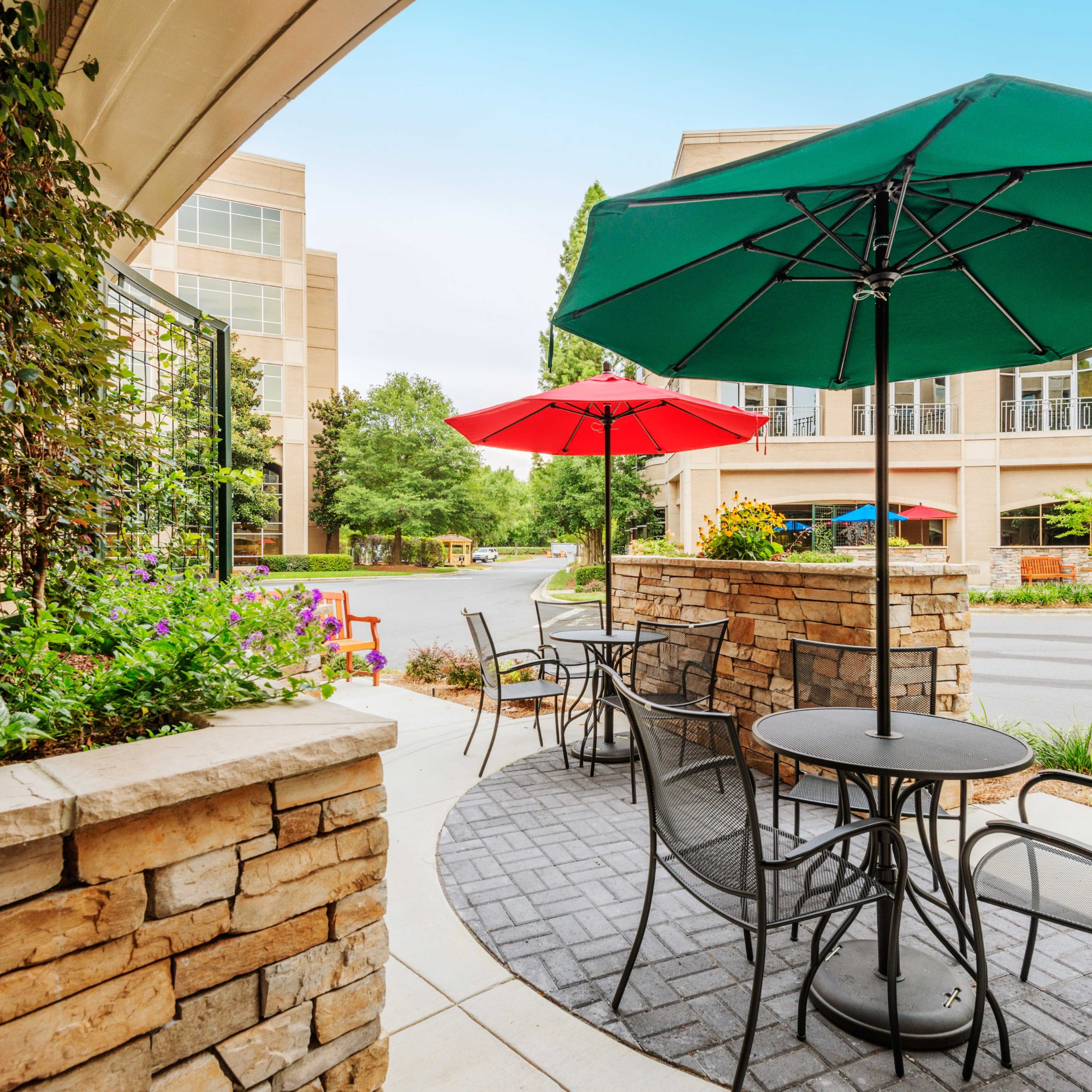 A cozy outdoor seating area features a round table and chairs under red and green umbrellas, surrounded by landscaped greenery and stonework.