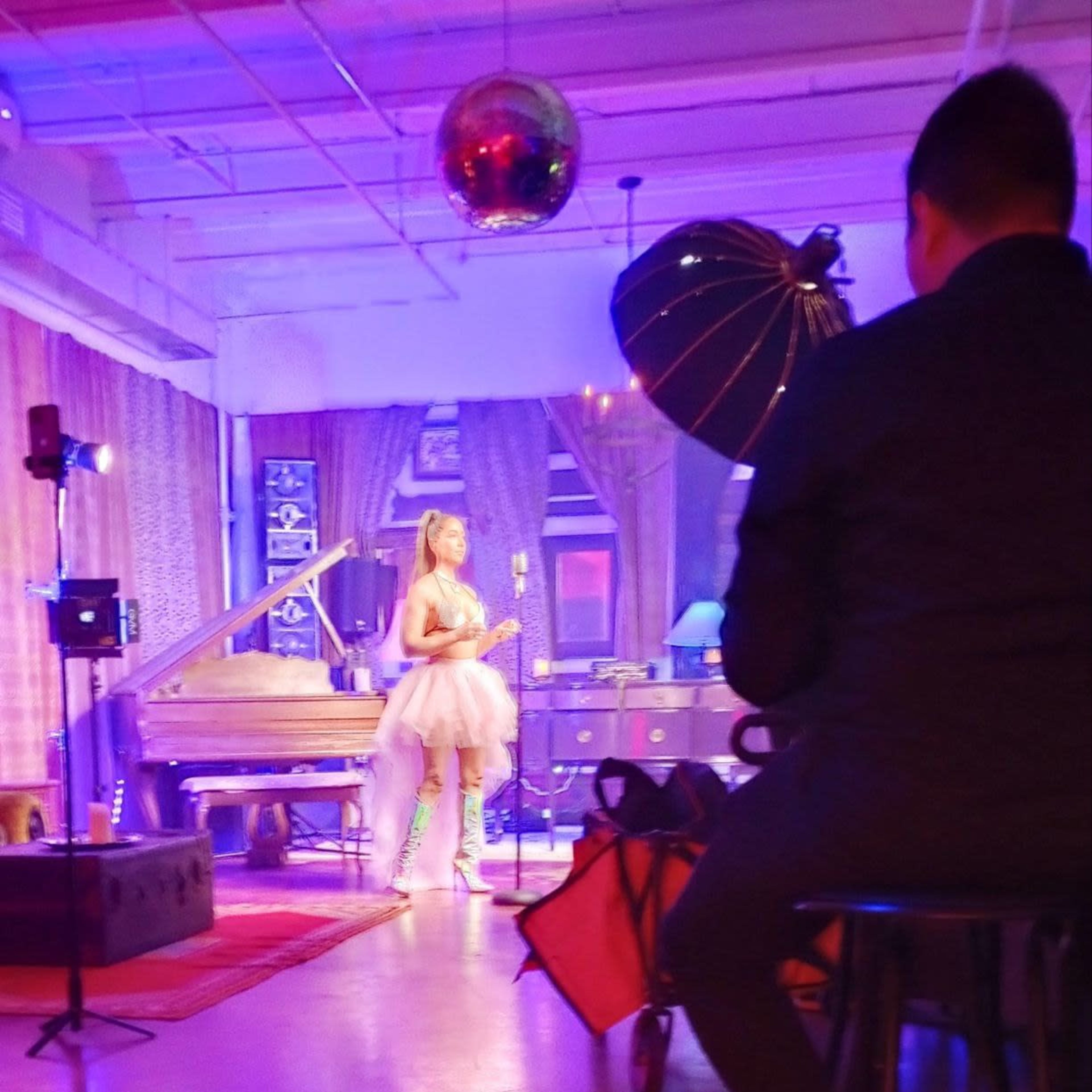 A woman in a tulle dress stands in a brightly lit studio with a grand piano, while a man adjusts lighting equipment.