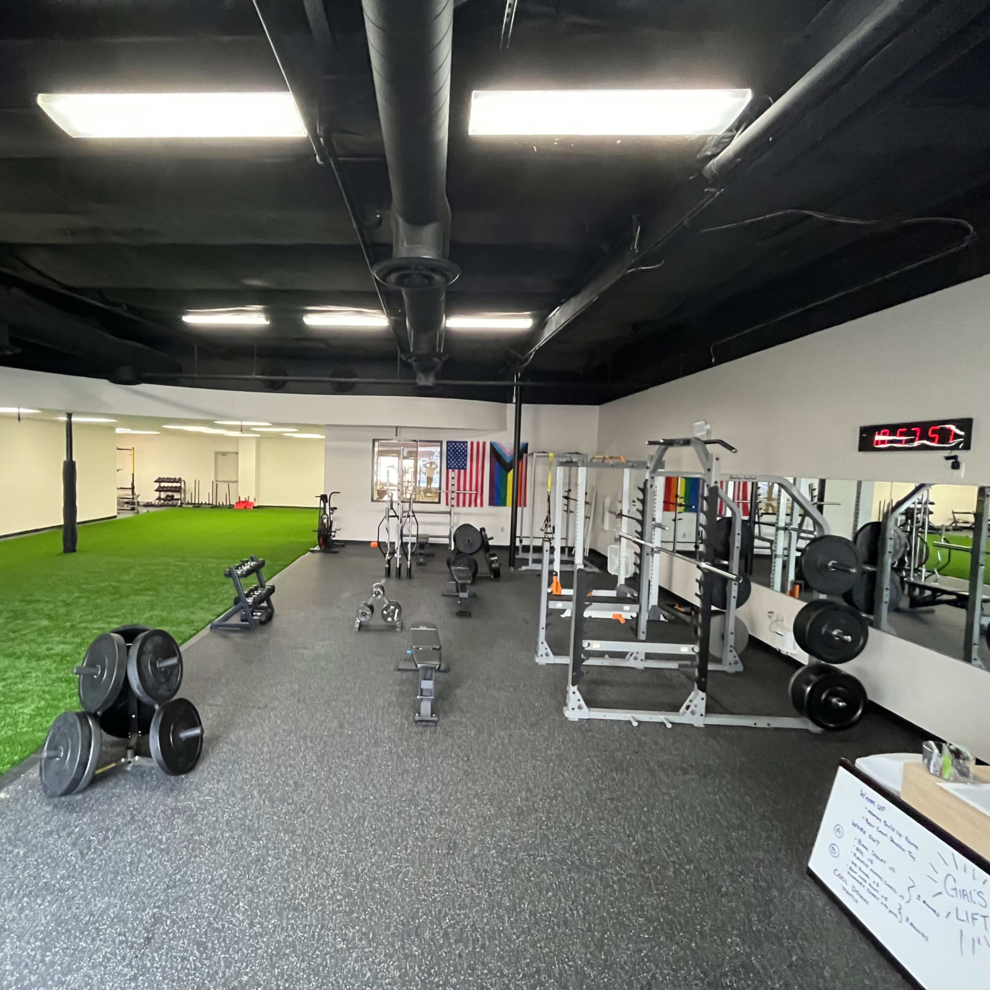 A gym interior with fitness equipment, including free weights and a squat rack, alongside a turf area for exercises.