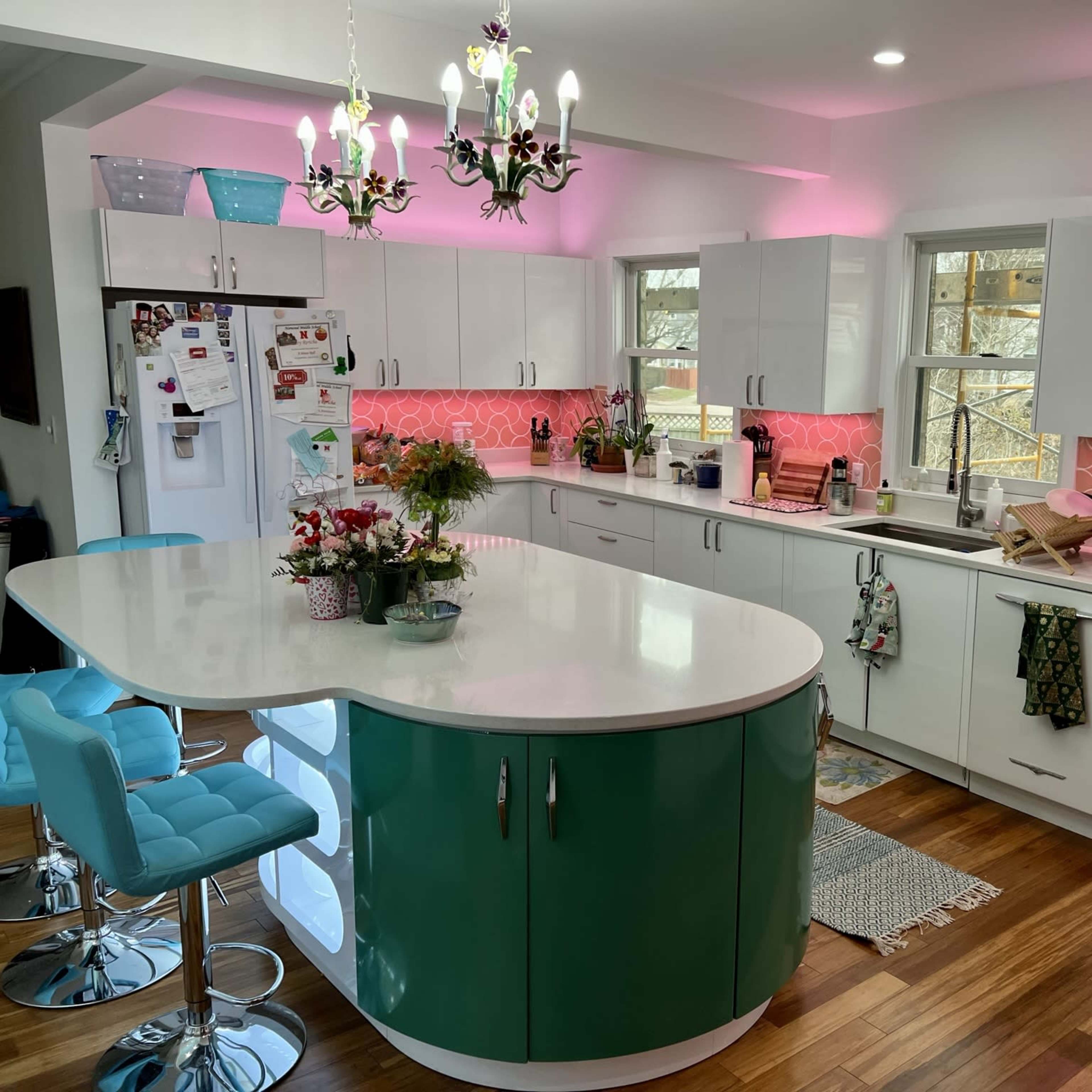 A modern kitchen with a large central island in teal, colorful LED lighting, and a variety of appliances and decorations along the countertops.