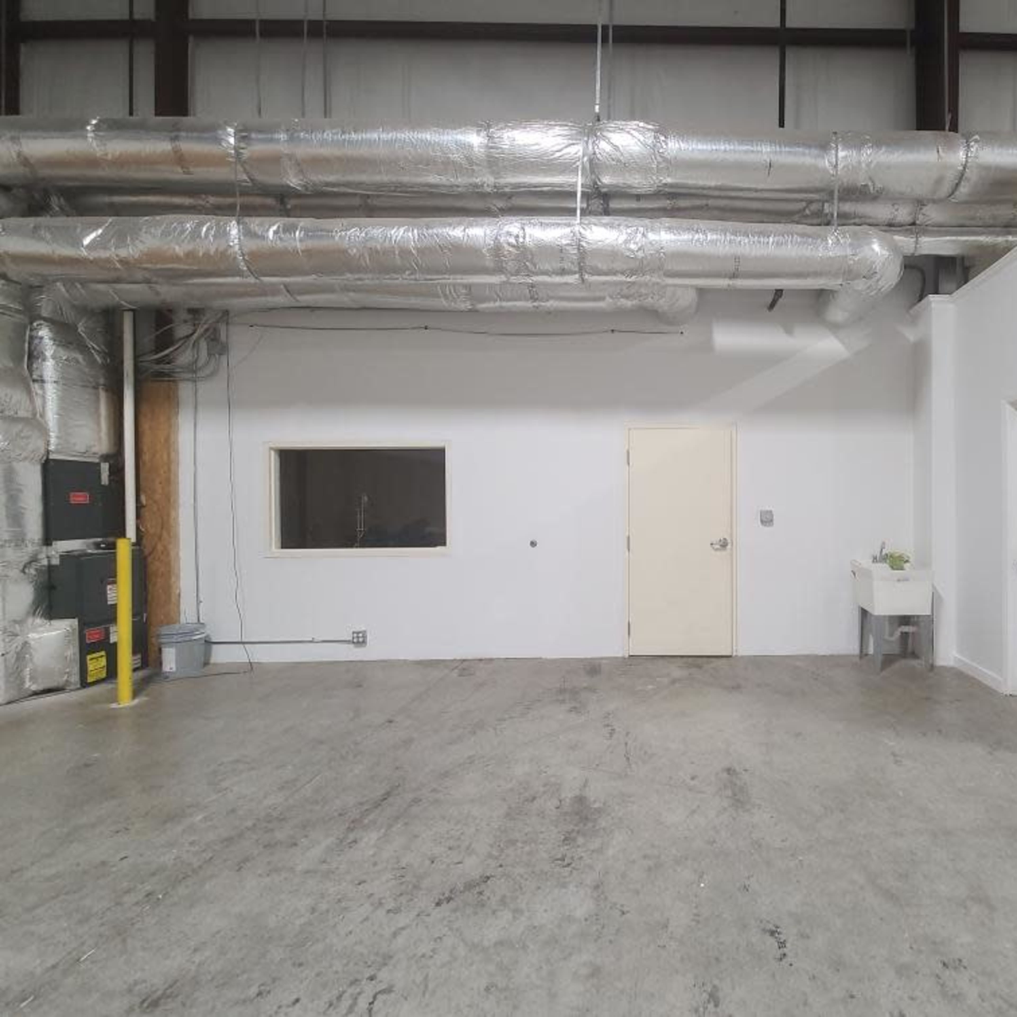 An empty industrial room with exposed metal ductwork, a door, and a small sink against a white wall.