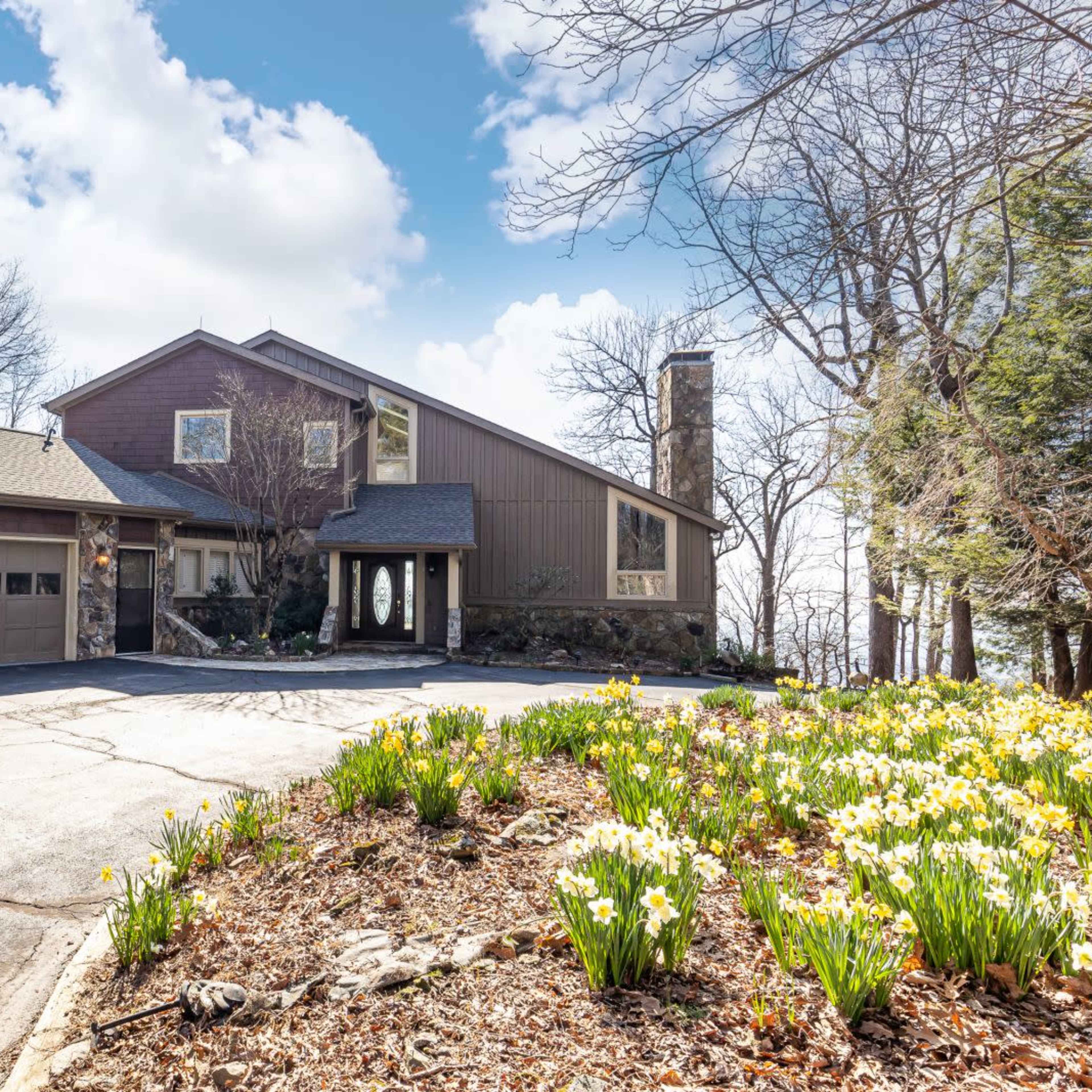 A modern, two-story house with a stone facade sits beside a paved driveway lined with blooming daffodils and surrounded by trees.