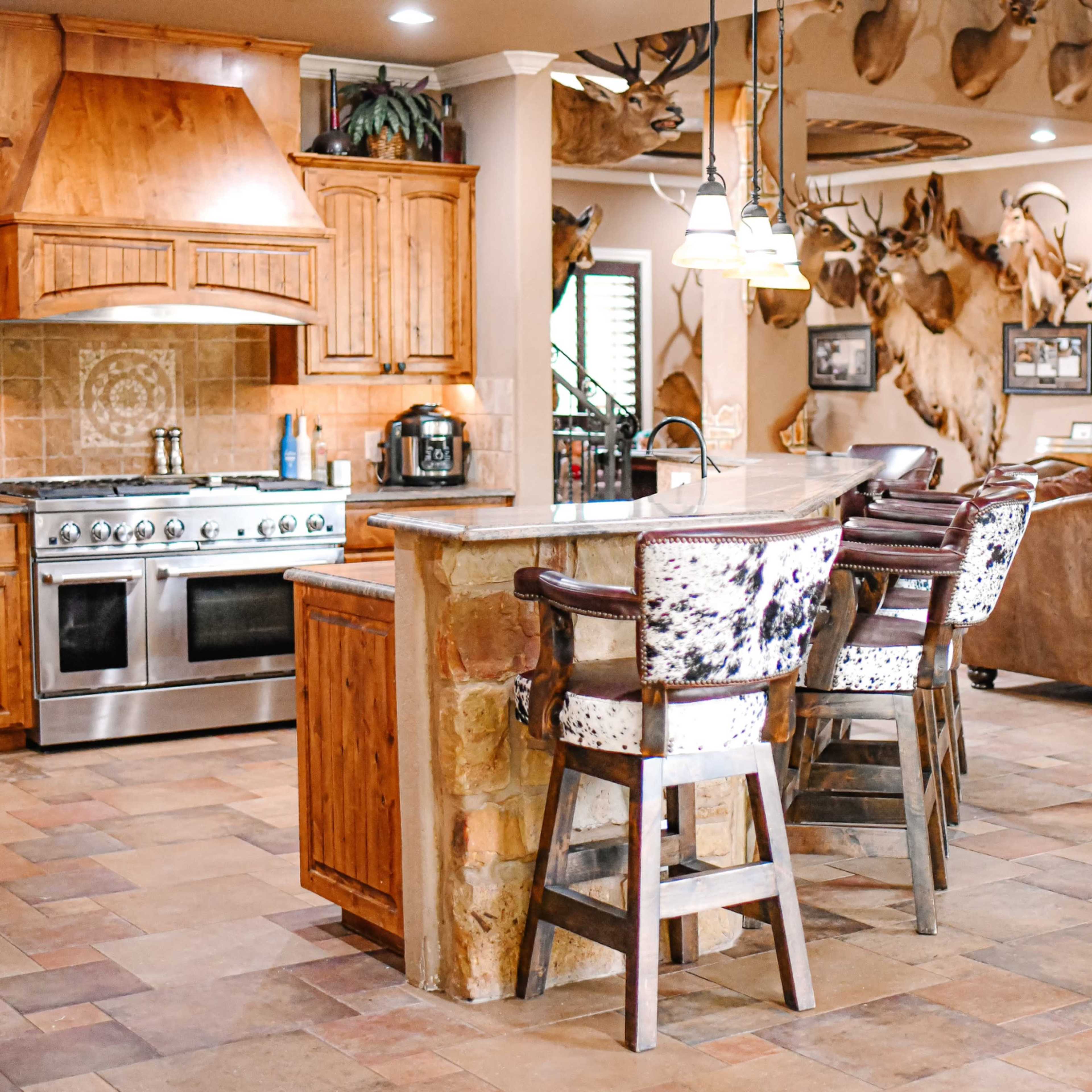 The kitchen features wooden cabinets, a large stove, and bar seating with patterned chairs, all set within a spacious room adorned with mounted animal heads on the walls.