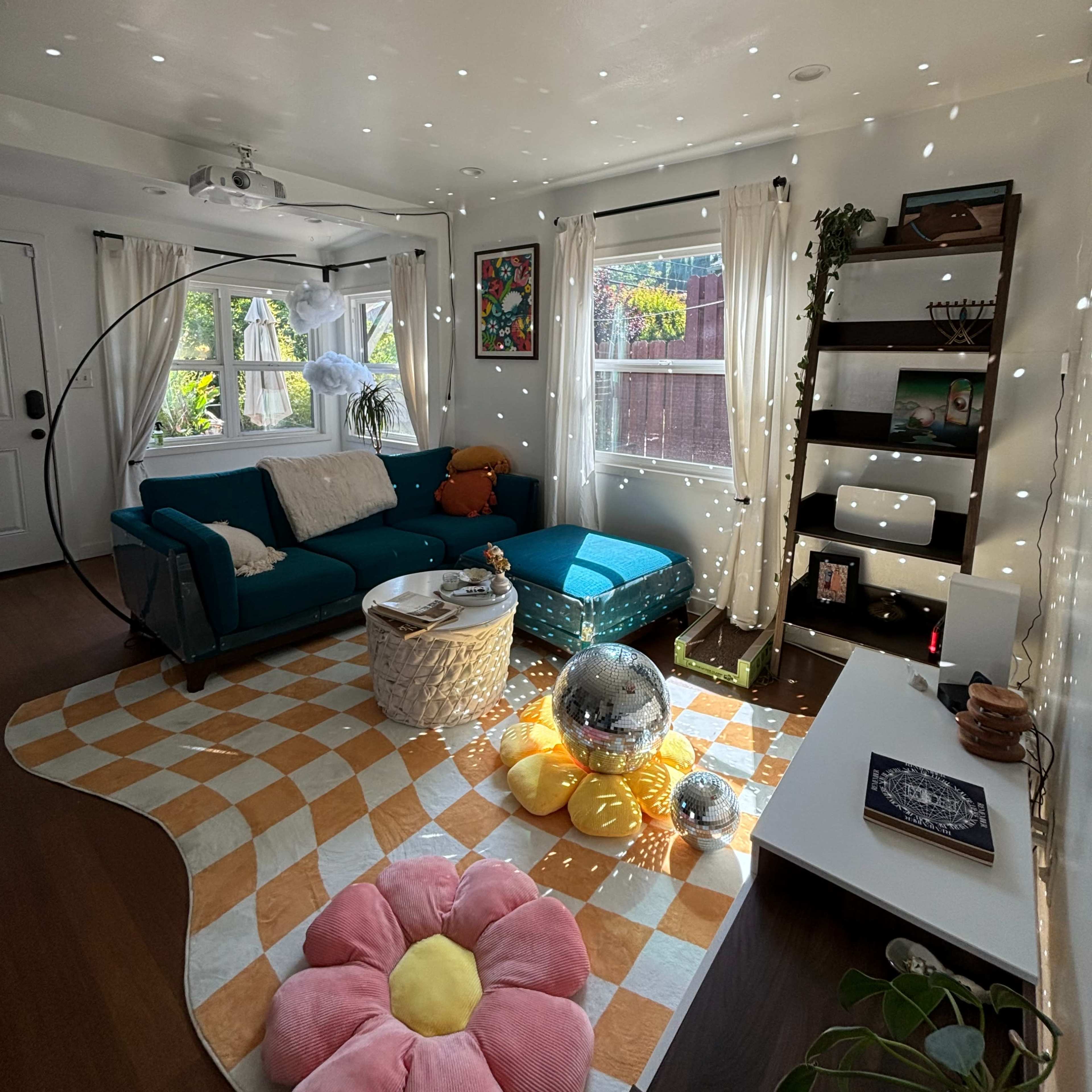 A brightly lit living room features a teal couch, a floral rug, and a disco ball reflecting light across the walls.