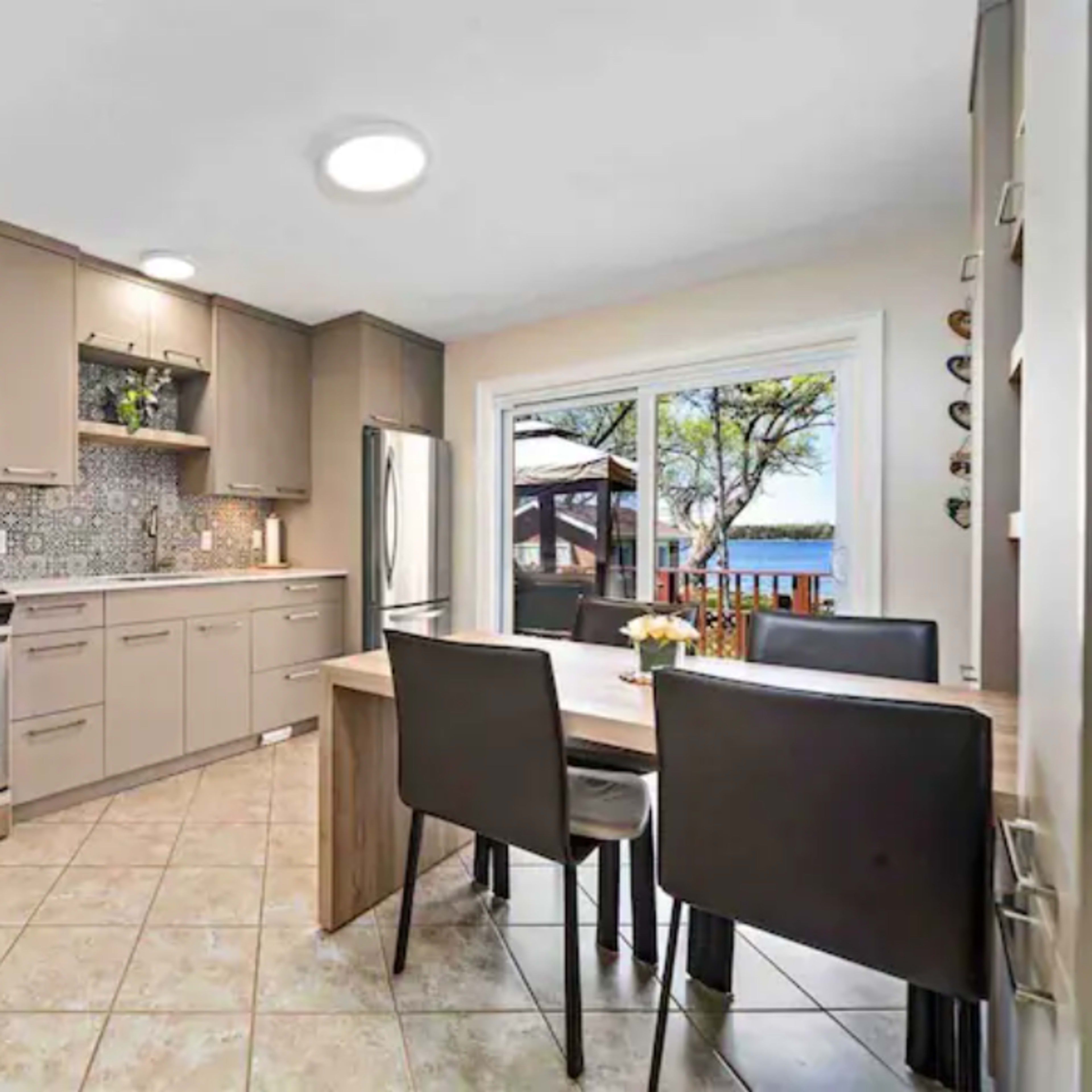 A modern kitchen with beige cabinets, a stainless steel oven and refrigerator, and a small dining table set against a backdrop of a view through large sliding glass doors.