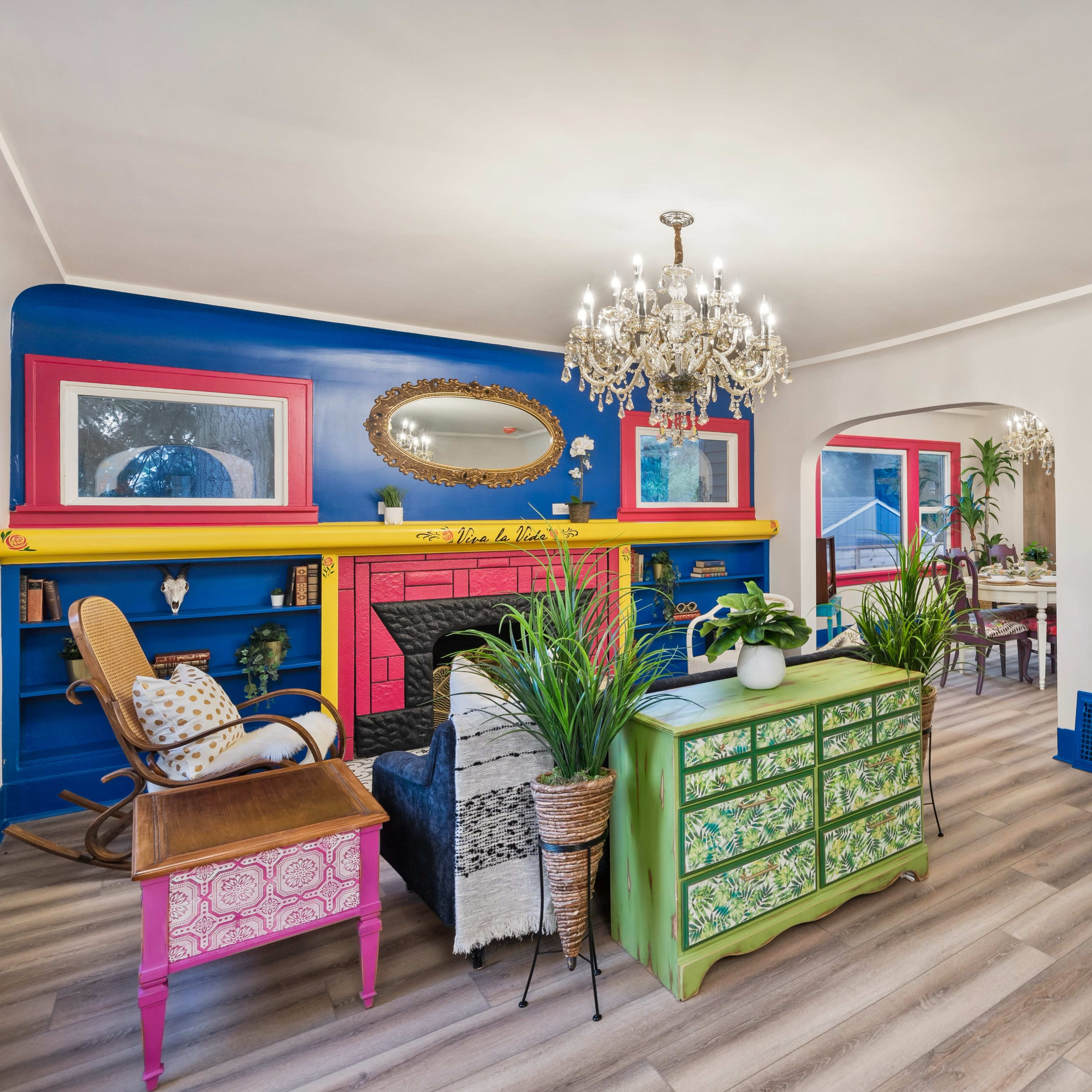A colorful living room features a vibrant blue and pink fireplace, a chandelier, decorative plants, and eclectic furniture.
